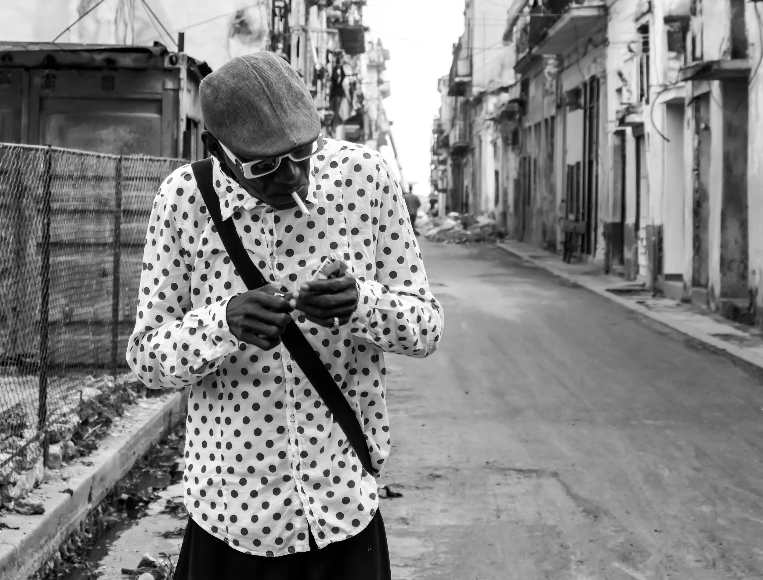 A man wearing a polka dot shirt, cap, and sunglasses leaning over in an urban alley, smoking a cigarette, looking at his phone, with dilapidated buildings and a fence in the background.