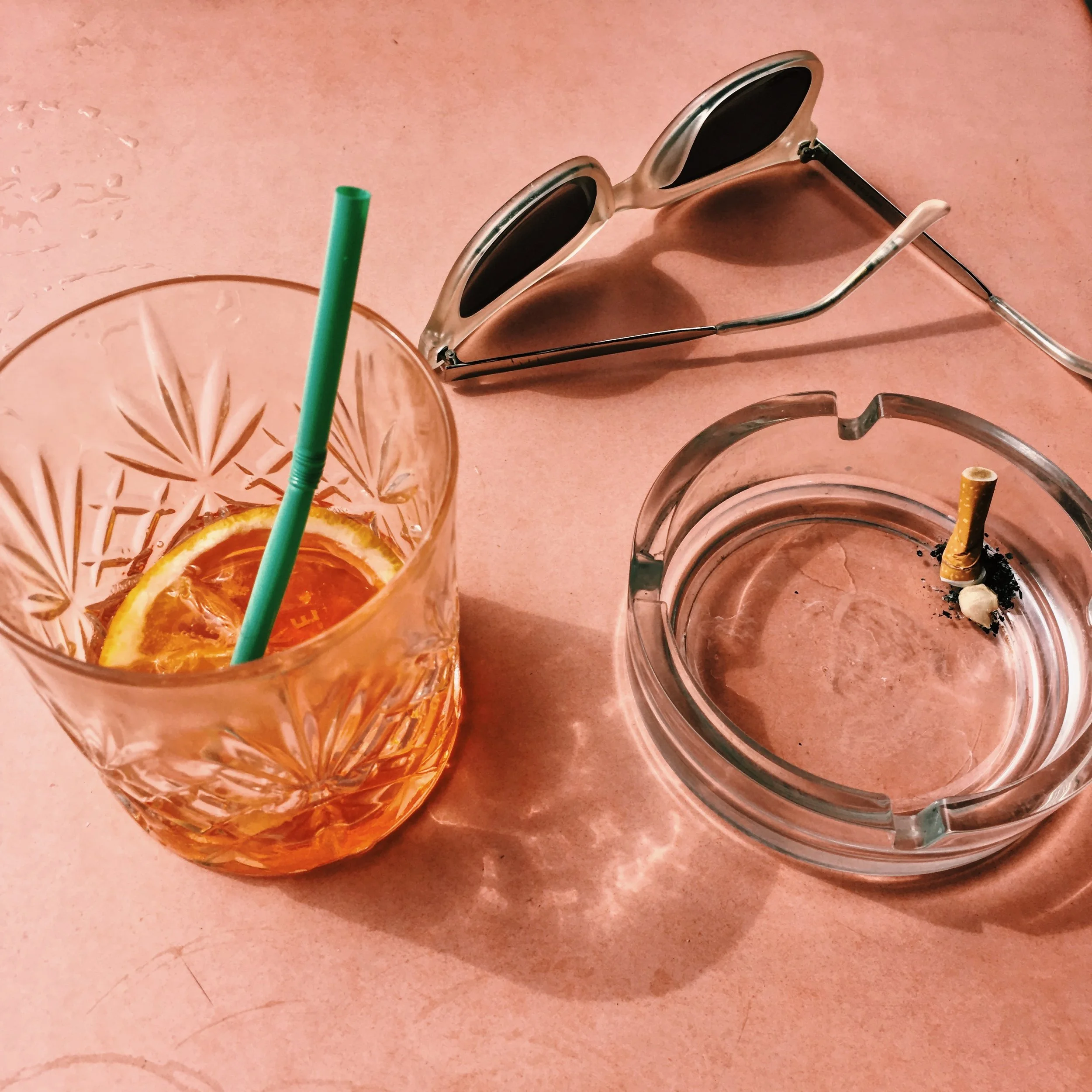 A glass with an orange slice and a green straw, beside a pair of sunglasses and an empty glass ashtray with a cigarette butt and black ashes, on a pink table.