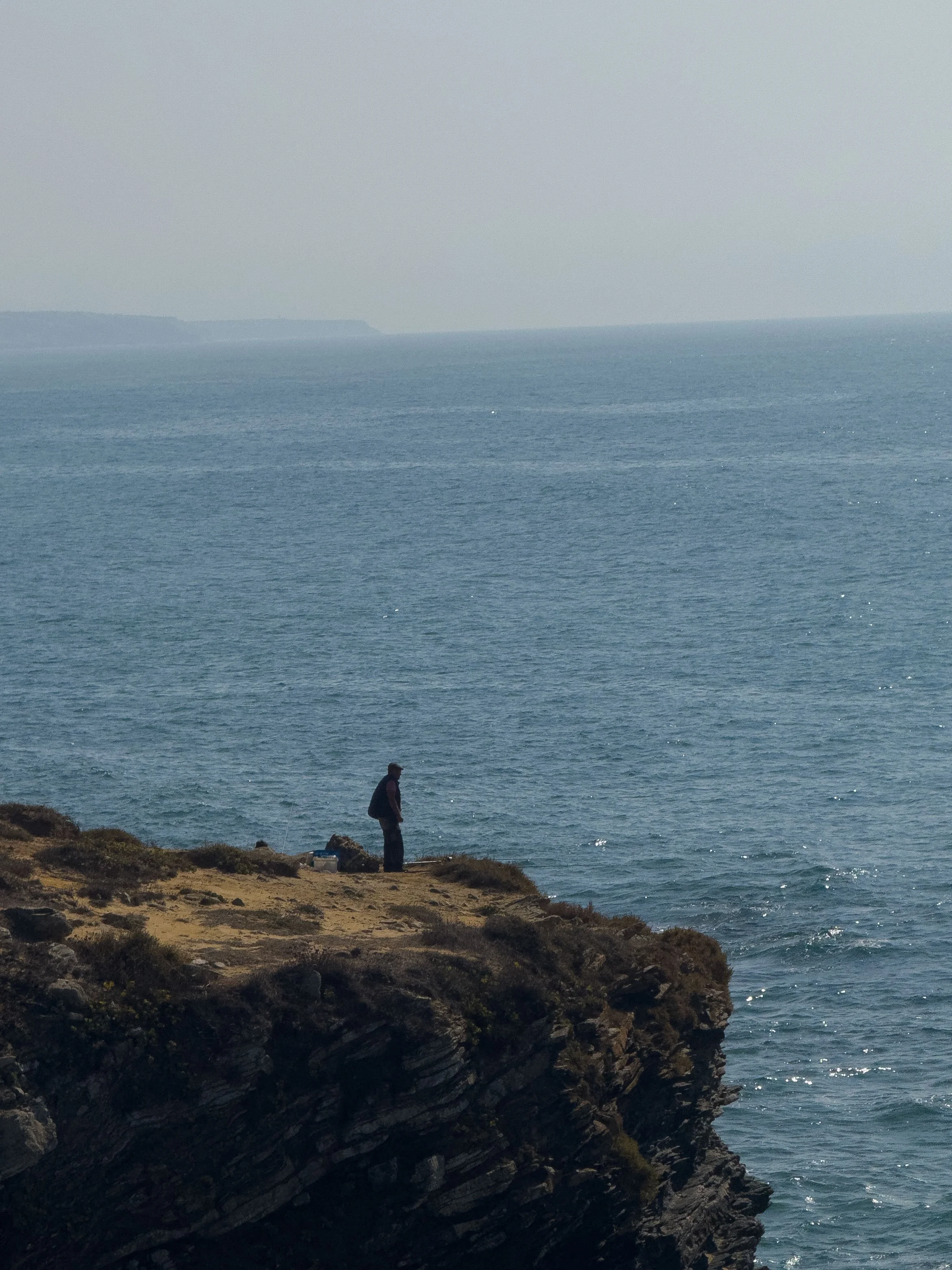 A person standing on a rocky cliff overlooking the ocean with a backpack.
