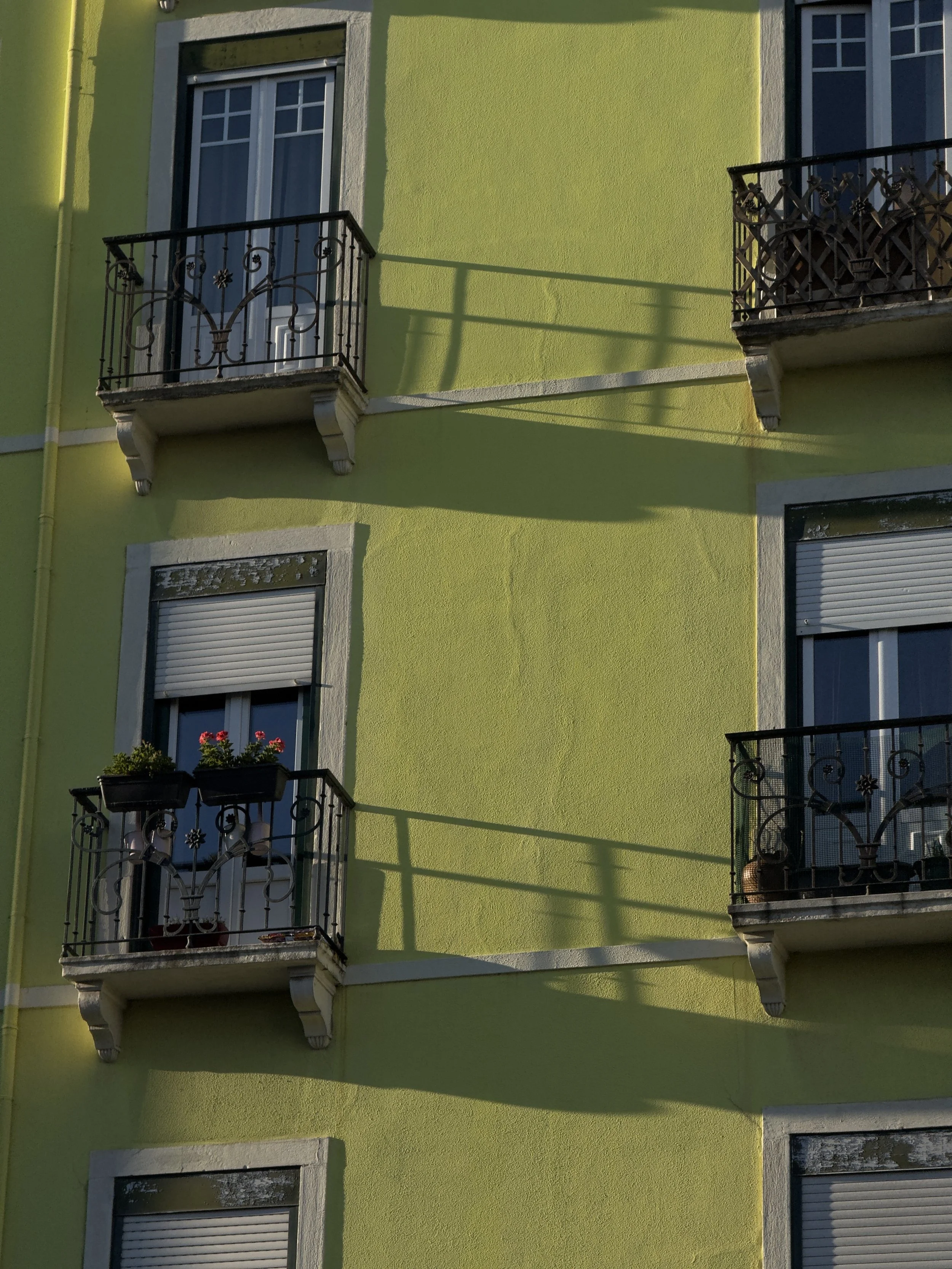 The image shows a three-story apartment building with a bright green exterior and white trim around the windows. There are balconies with decorative black metal railings, some with potted plants and flower boxes. The sunlight casts shadows of the bal