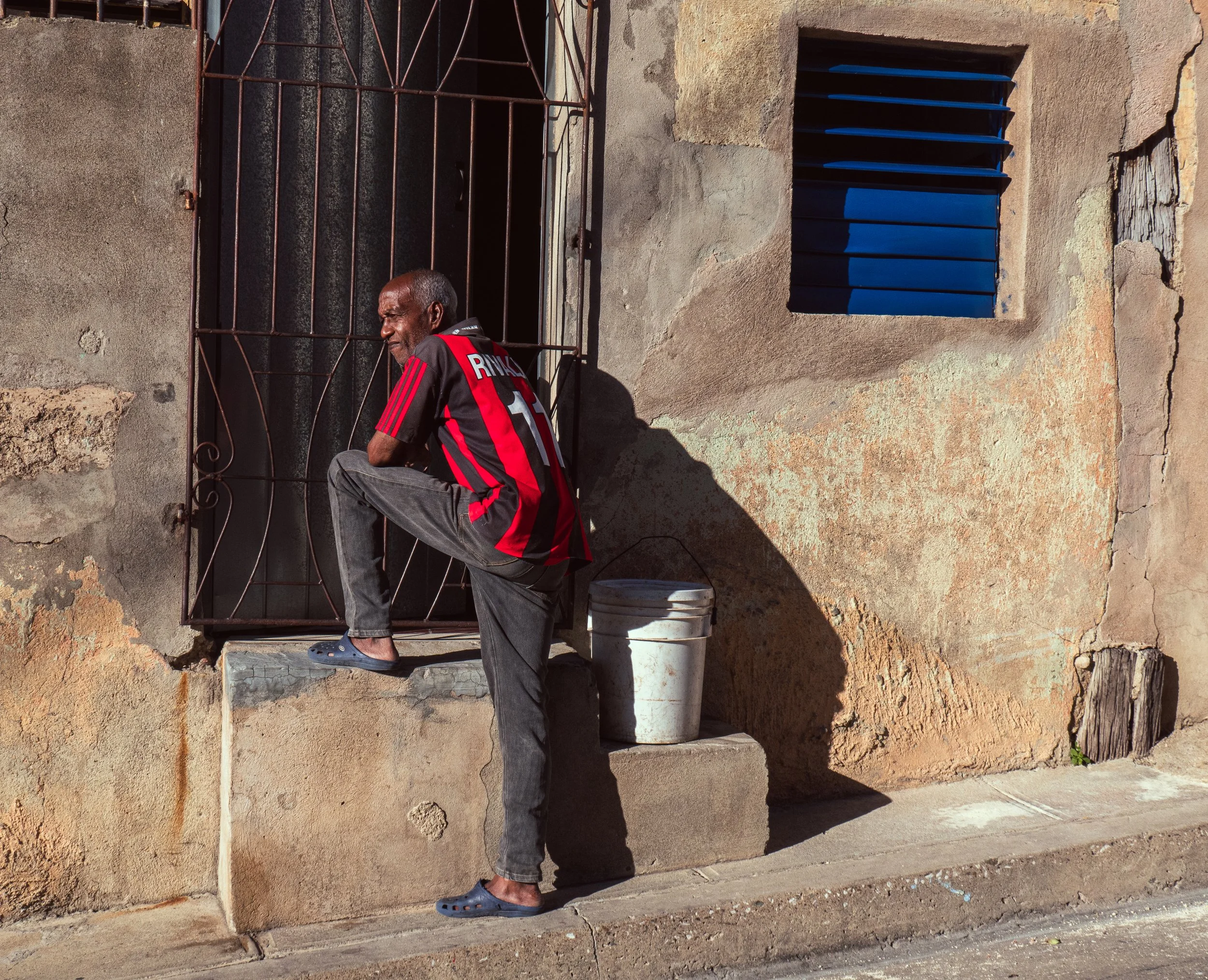 An older man wearing a black and red soccer jersey and gray jeans standing on a small concrete ledge outside an old building with a rough, weathered wall and a blue window with metal bars, soaking up the sun.