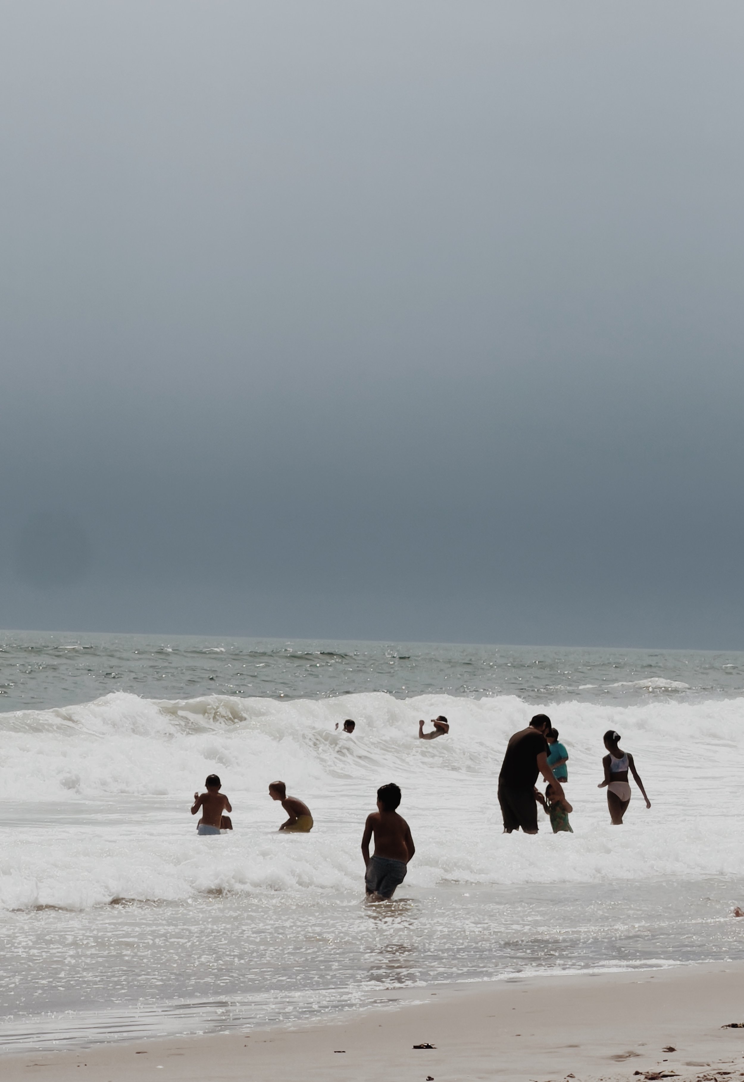 People enjoying a day at the beach, some swimming and others standing or walking in the surf, with an overcast sky.