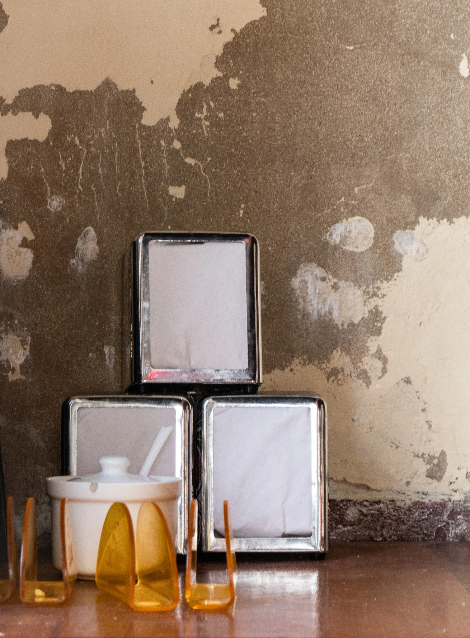 Three empty metal napkin holders stacked on a wooden surface next to a cup of cream with a straw, against a distressed wall.