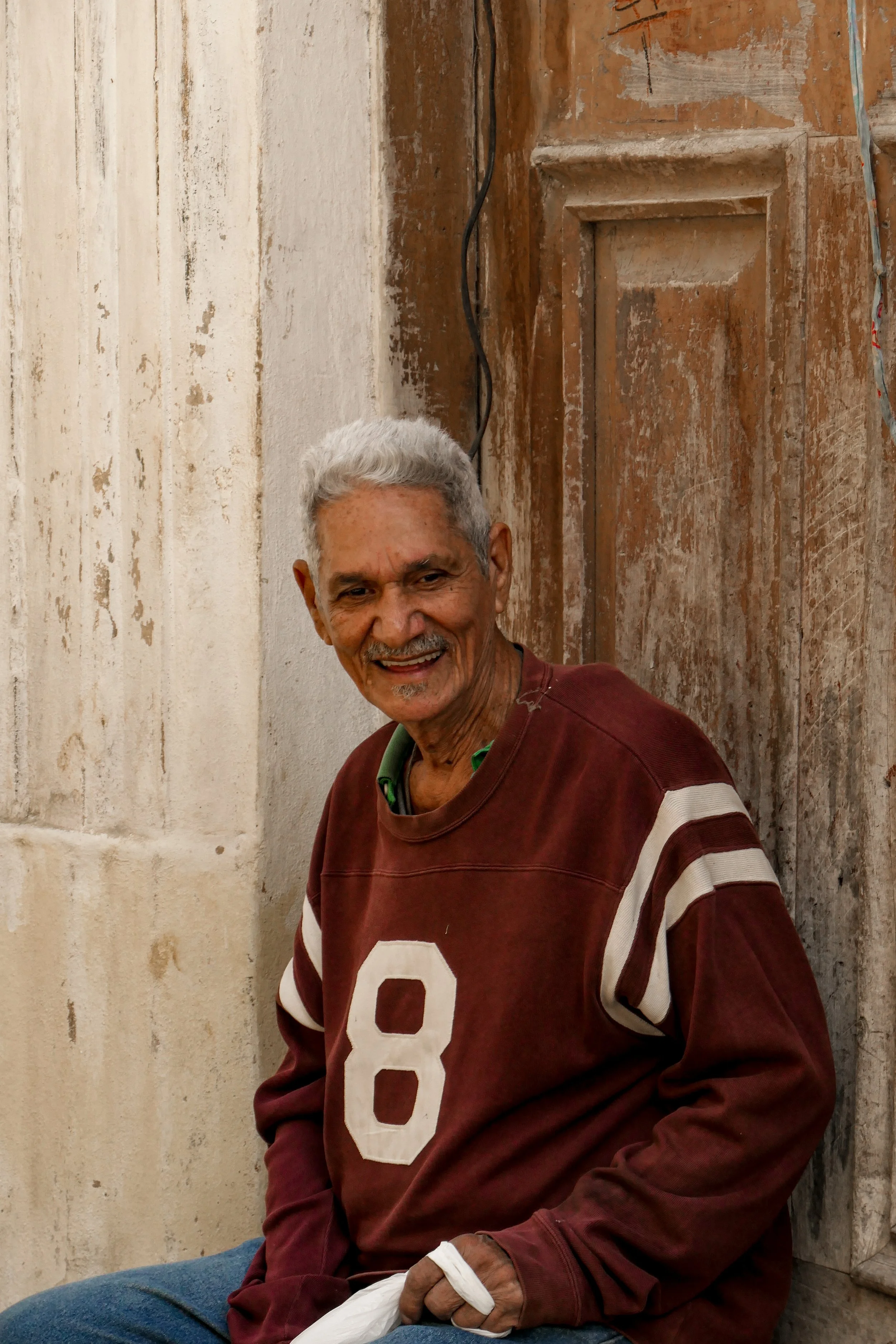 An elderly man with gray hair and a warm smile, wearing a maroon sweatshirt with the number 8, sitting near a textured wall and weathered wooden door.