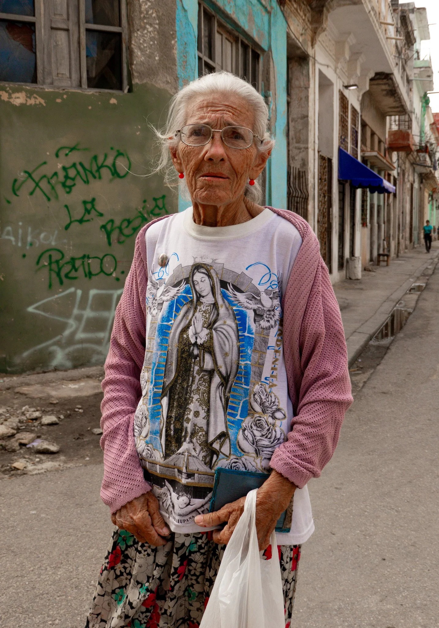 Elderly woman standing on a city street with buildings and graffiti in the background, wearing glasses, a religious-themed graphic T-shirt, pink sweater, and patterned skirt, holding a plastic bag and a smartphone.
