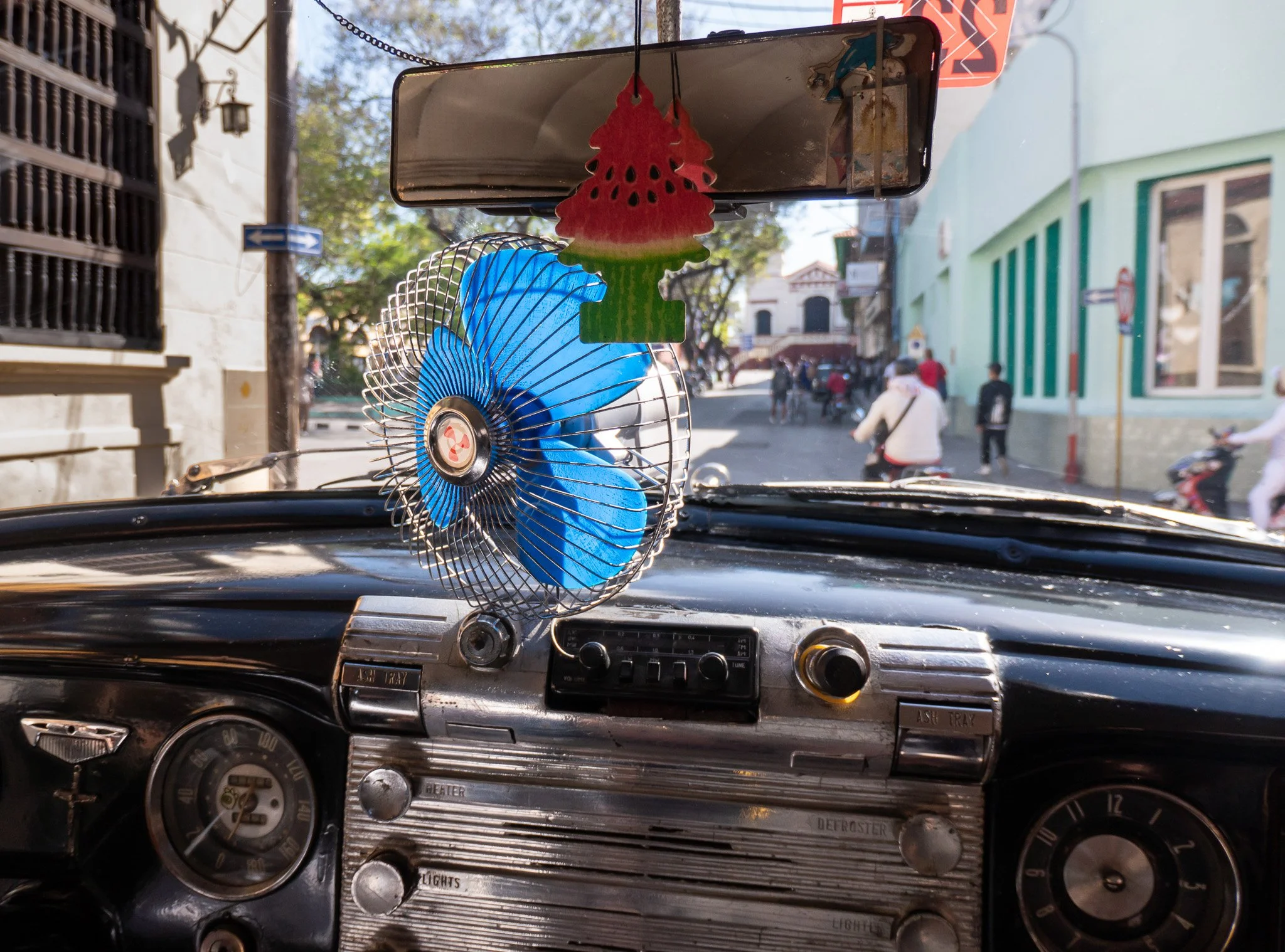 View from the dashboard of an old car looking out onto a street with pedestrians and buildings, with a small blue fan and colorful air fresheners hanging from the rearview mirror.
