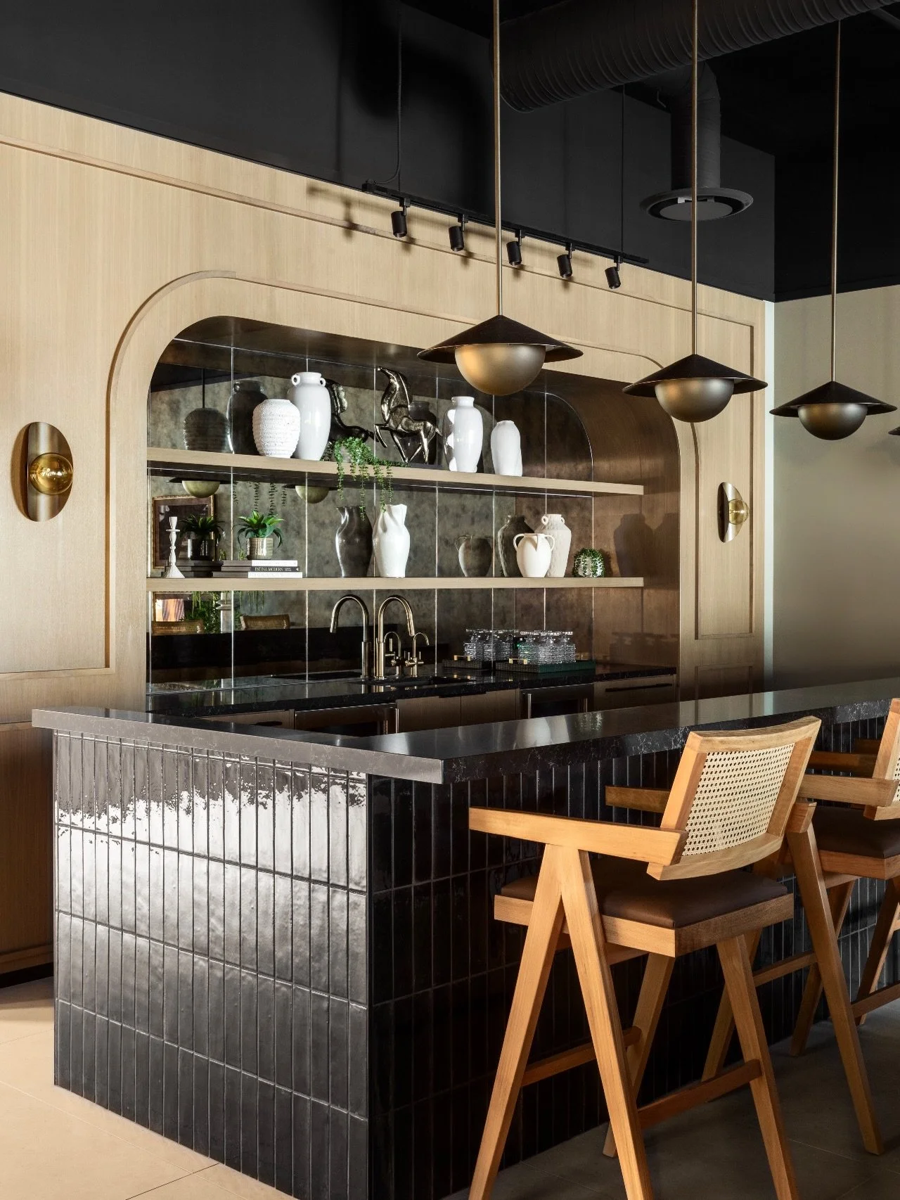 The Bar. A seamless and bold moment at #AVDCalgaryHouse. The tile-clad island acts as a dark anchor against the softly curved light-wood millwork, layered with inset floating cabinetry, antique mirror for added drama, and striking custom light fixtur