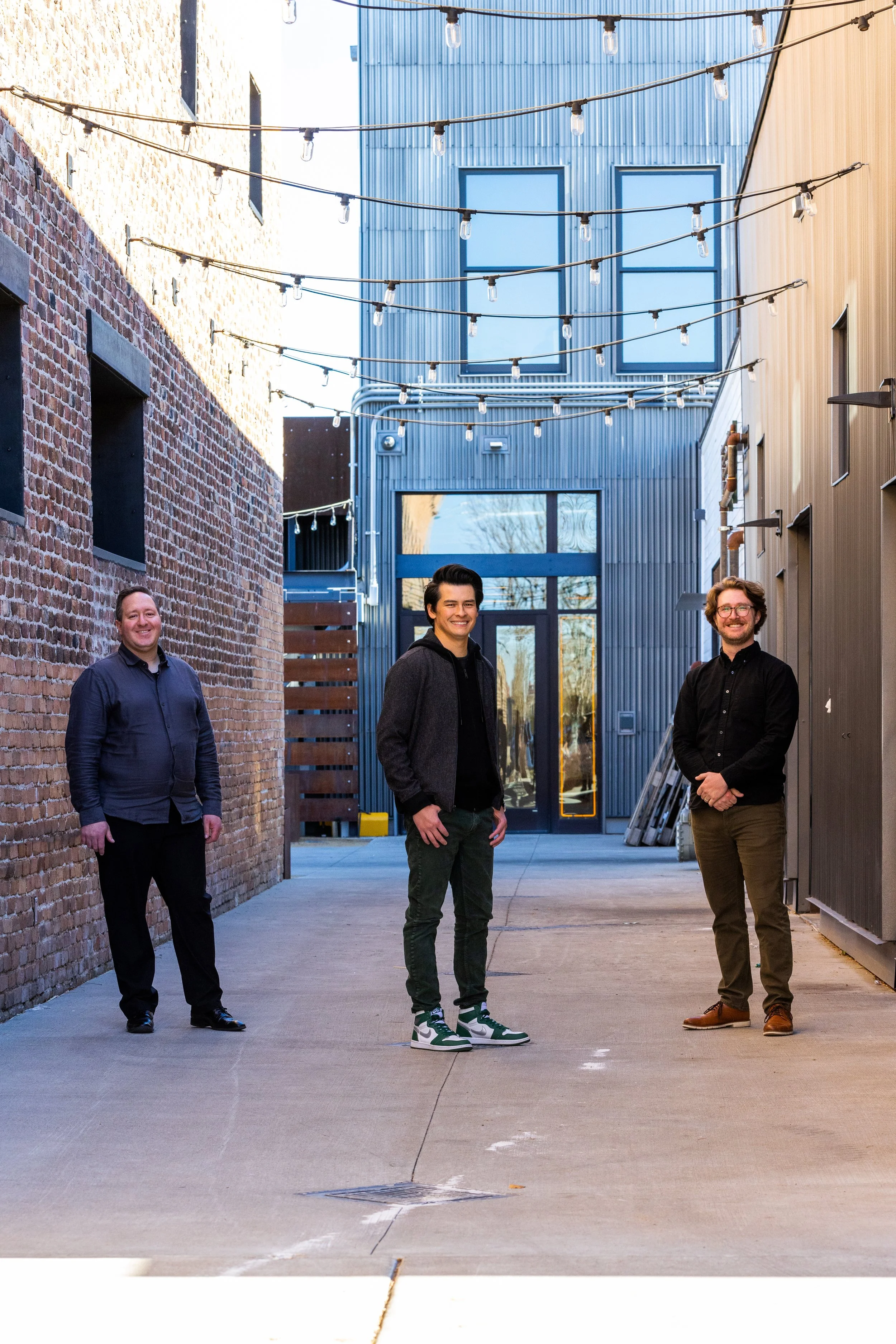 Four men standing in an alleyway between brick and metal buildings, decorated with string lights overhead, with a partially reflected sky and tree in the background.