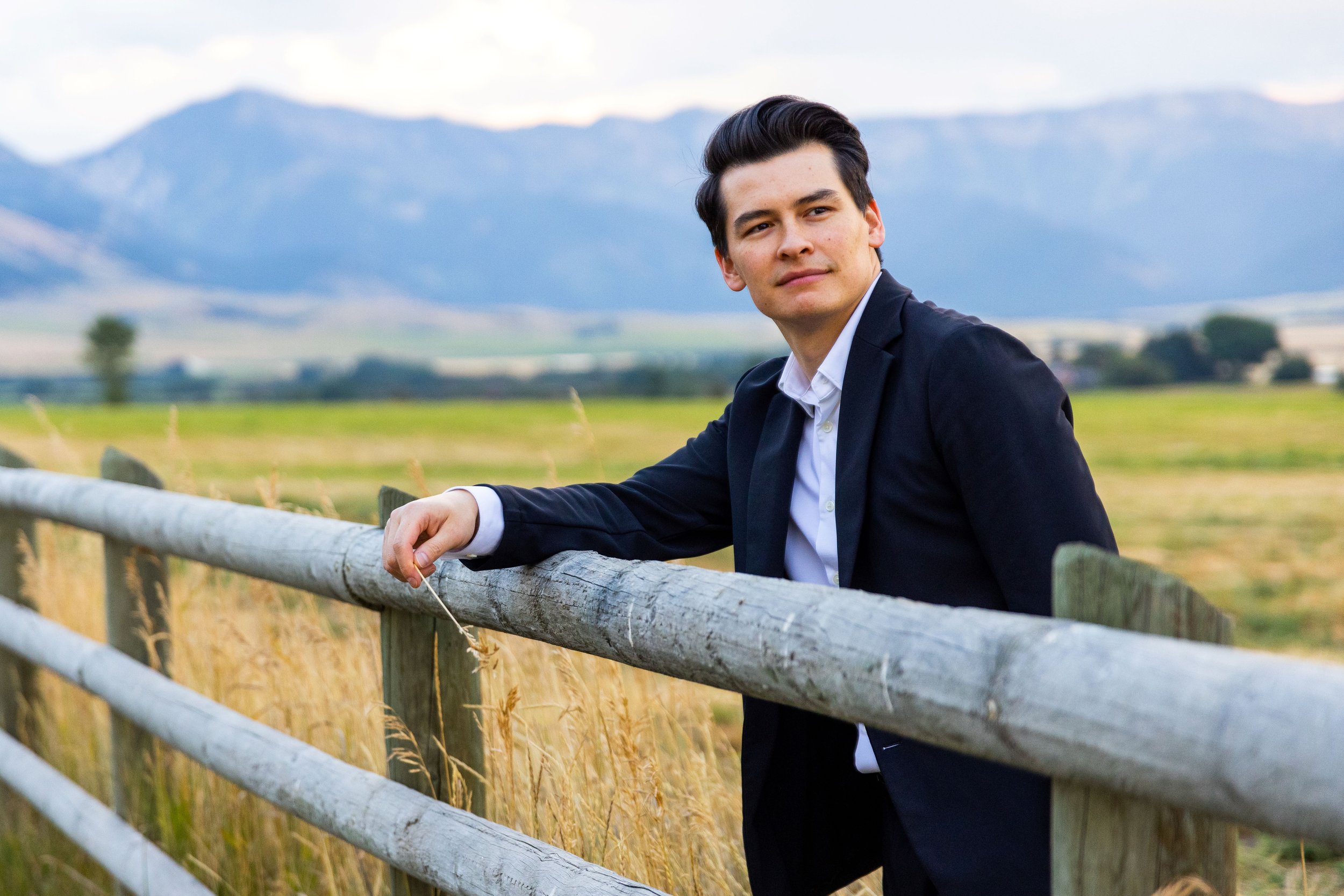 A young man in a field, leaning on a wooden post fence. He is staring off into the distance and the mountains are behind him