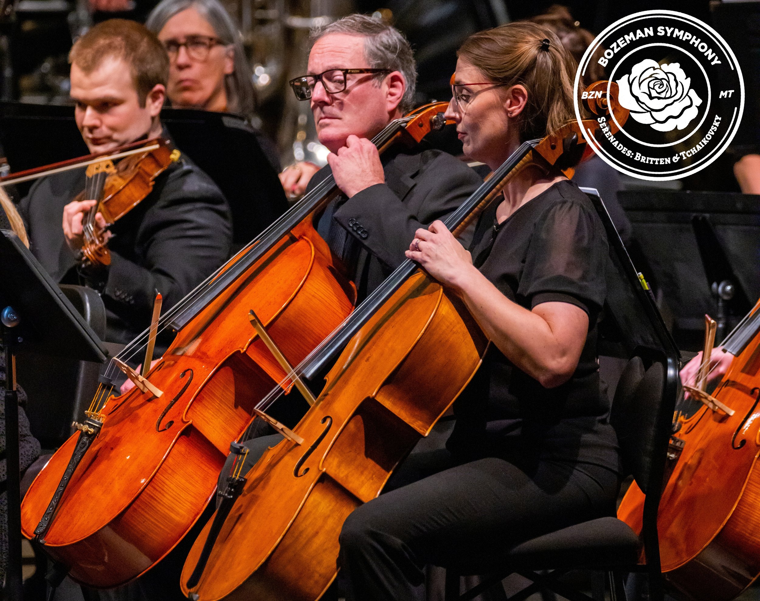 An orchestra performance, featuring musicians playing violins in the foreground and a cellist, a clarinetist, and a tuba player in the background, all dressed in black, on a dimly lit stage.