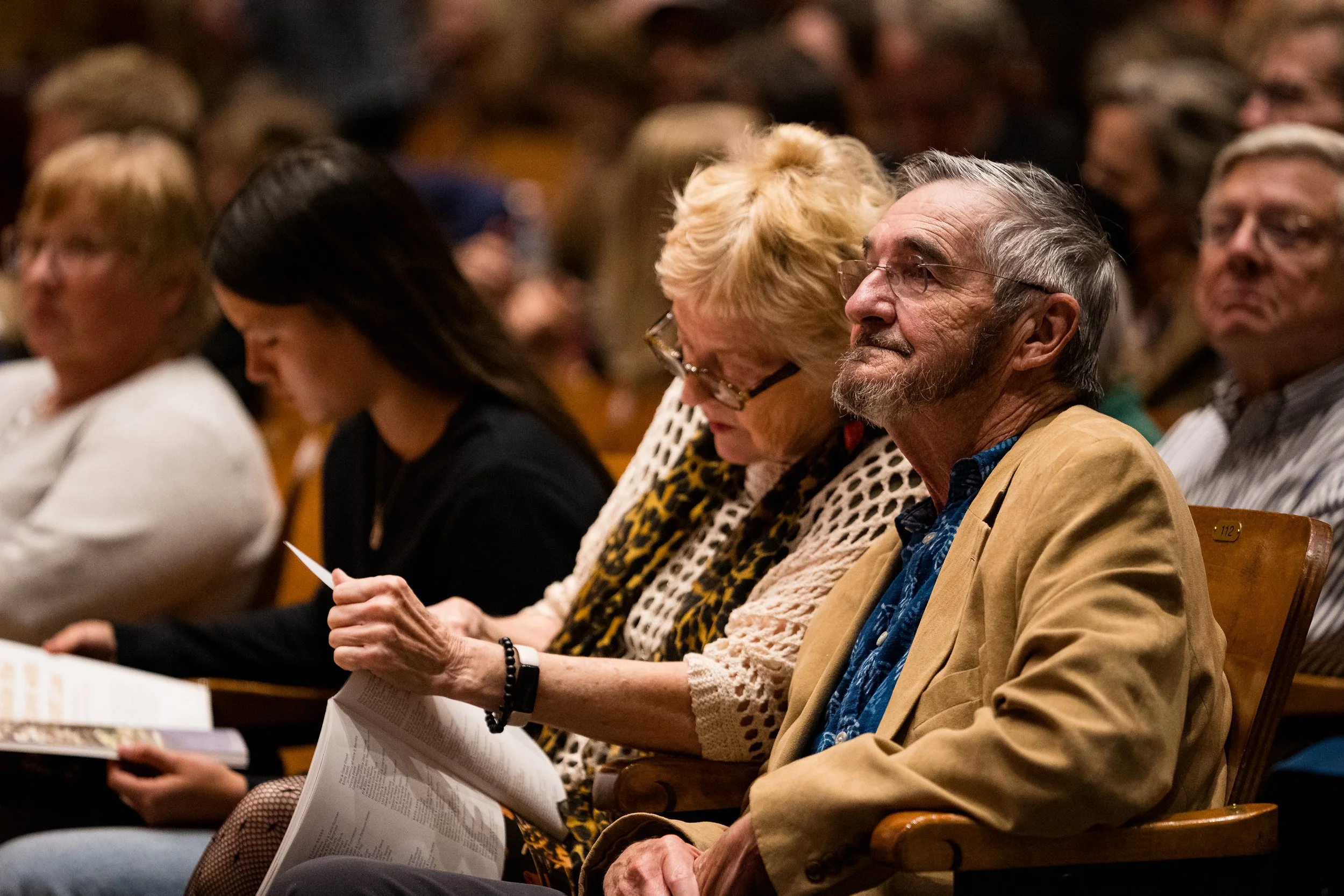 An older couple is sitting in the audience. The man is looking up and slightly smiling at the stage and the woman is looking intently at the concert program