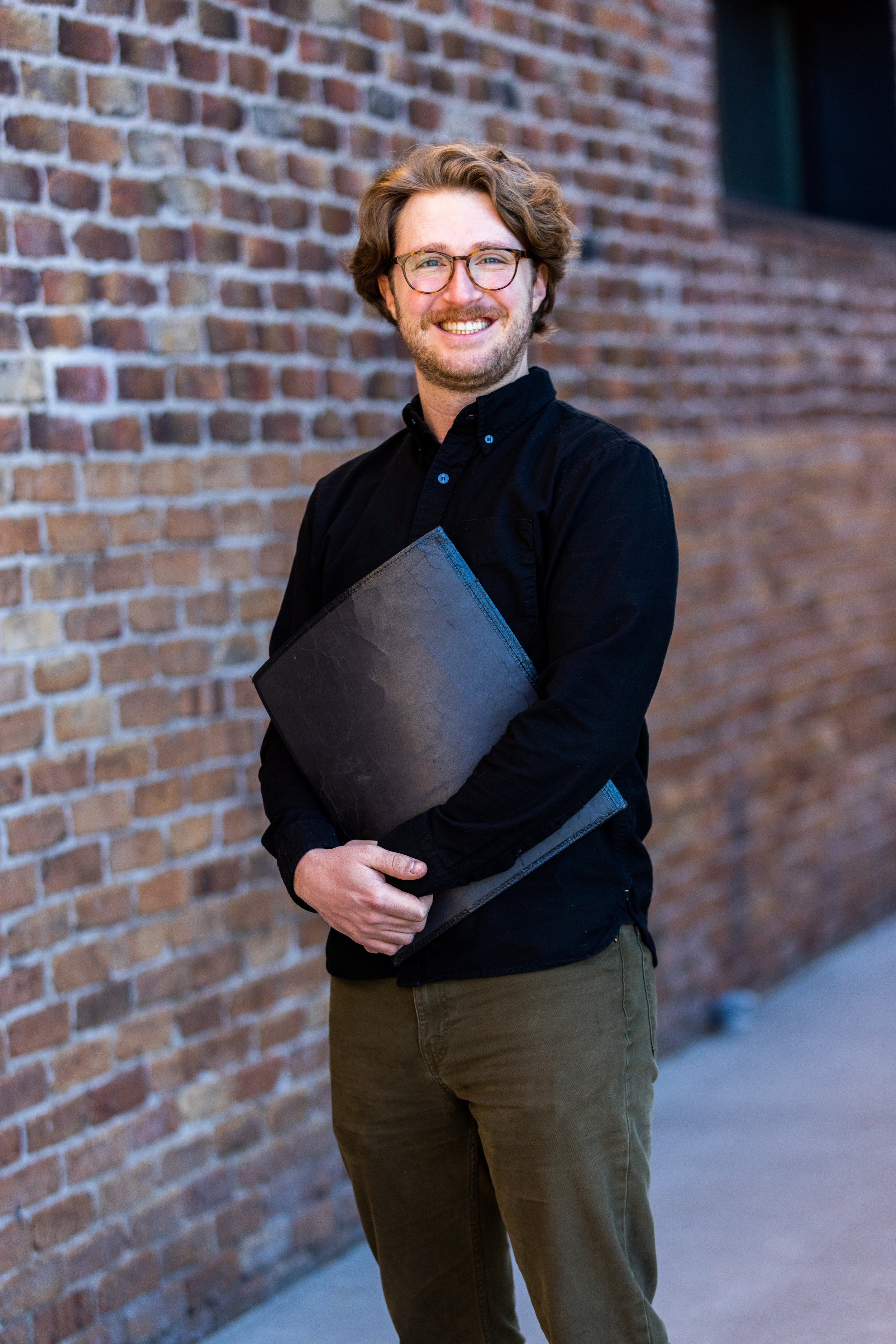 A young man with glasses, wavy brown hair, and a beard, smiling and holding a closed choir folder, stands in front of a brick wall.