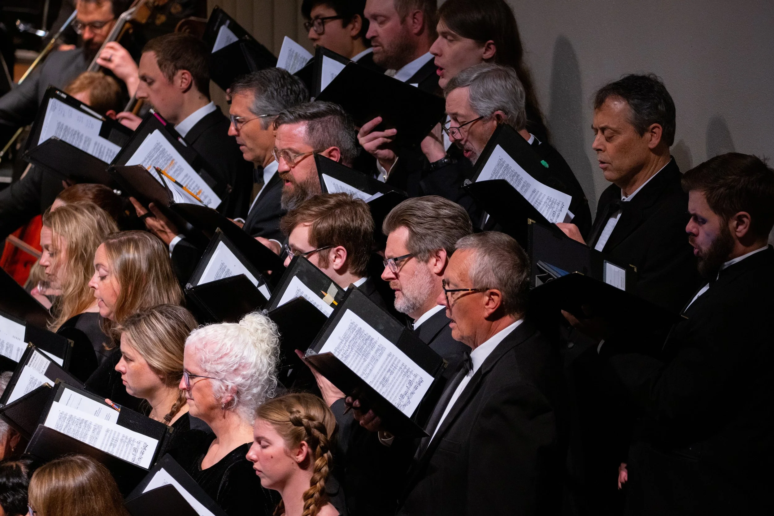 A group of musicians, including men and women of various ages, are performing with sheet music in front of them. They are dressed in formal black attire and are holding musical instruments or singing, suggesting they are part of a choir or orchestra, attending a concert or formal event.