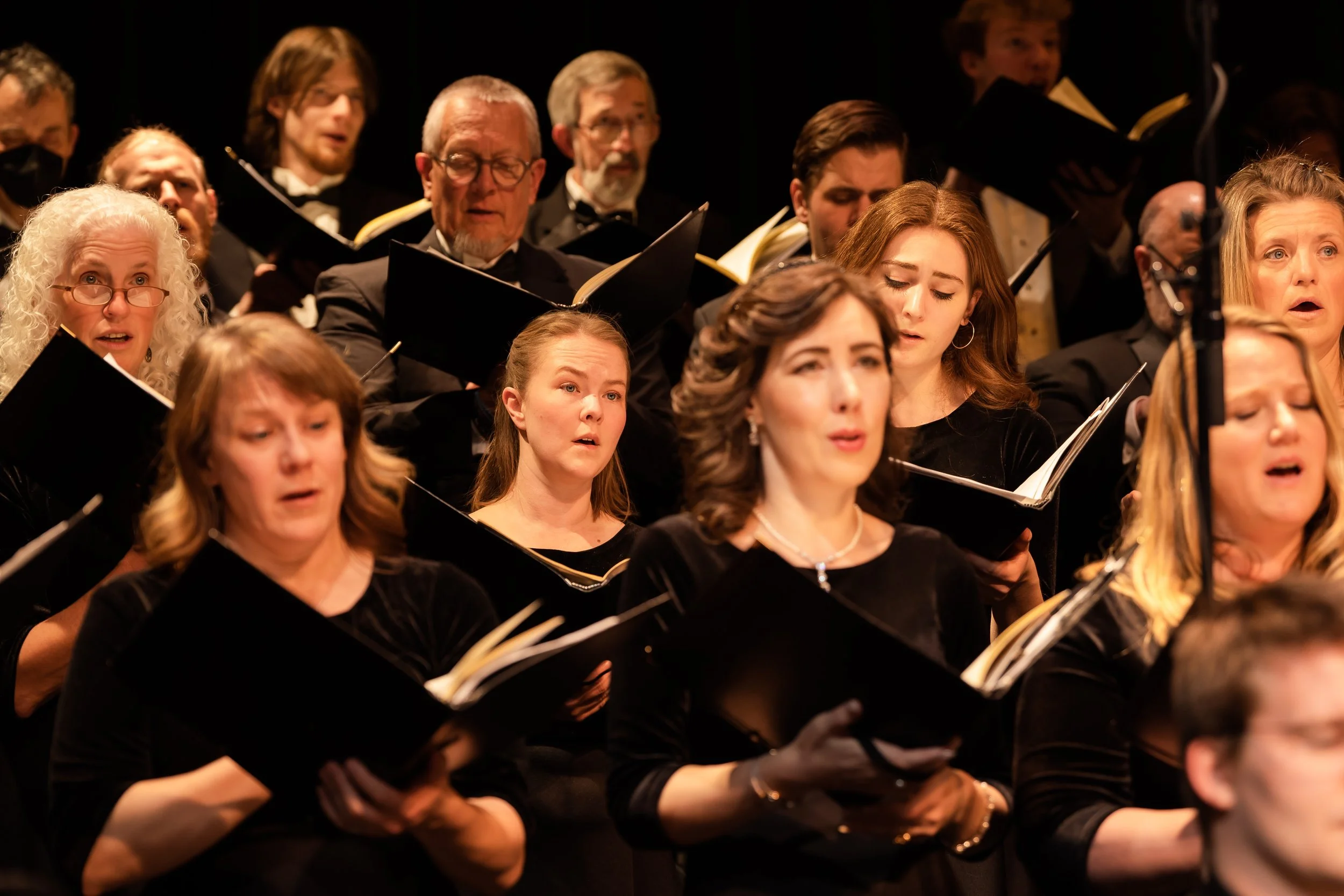 A group of choir singers performing, holding sheet music, dressed in black outfits in a concert setting.