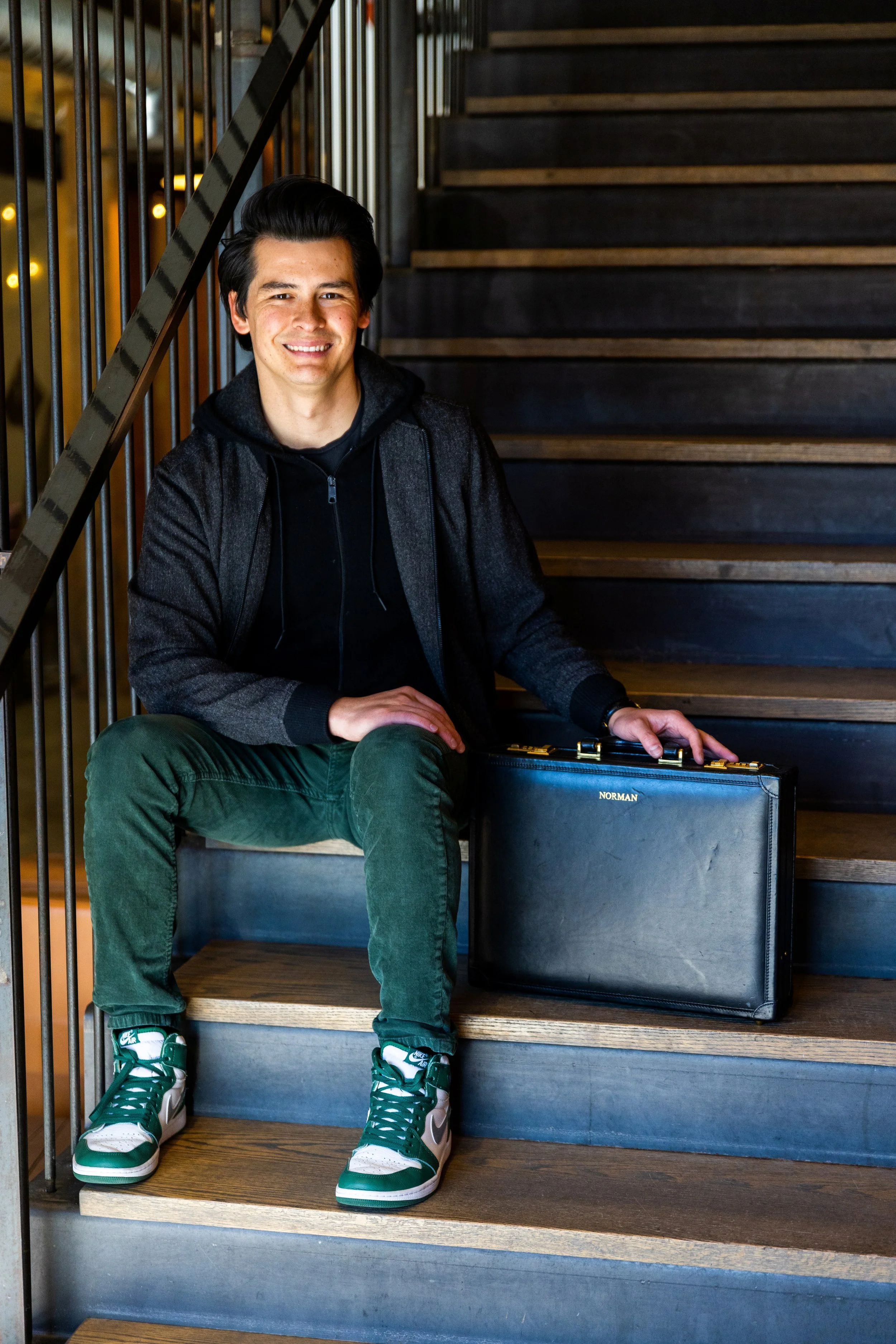 Young man sitting on wooden stairs, smiling, with a black briefcase labeled 'Norman' and a backpack on his lap, dressed in a black hoodie, gray jacket, green pants, and green and white sneakers.