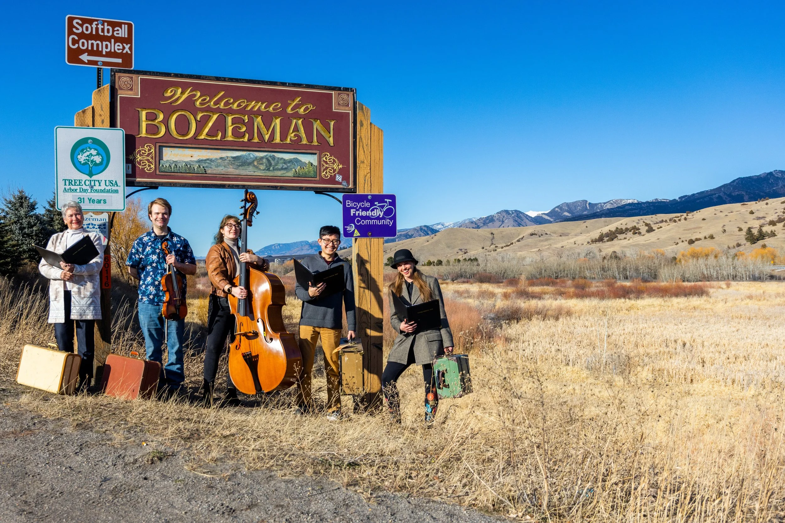 A group of five musicians standing outdoors in front of a 'Welcome to Bozeman' sign with mountains in the background. They are holding musical instruments and sheet music, with luggage and cases on the ground.