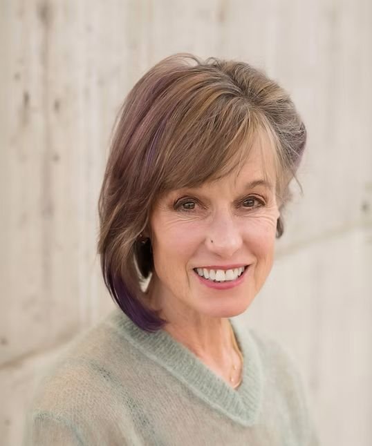 A middle-aged woman with short light brown hair, smiling, wearing a light-colored sweater, standing against a wooden wall.
