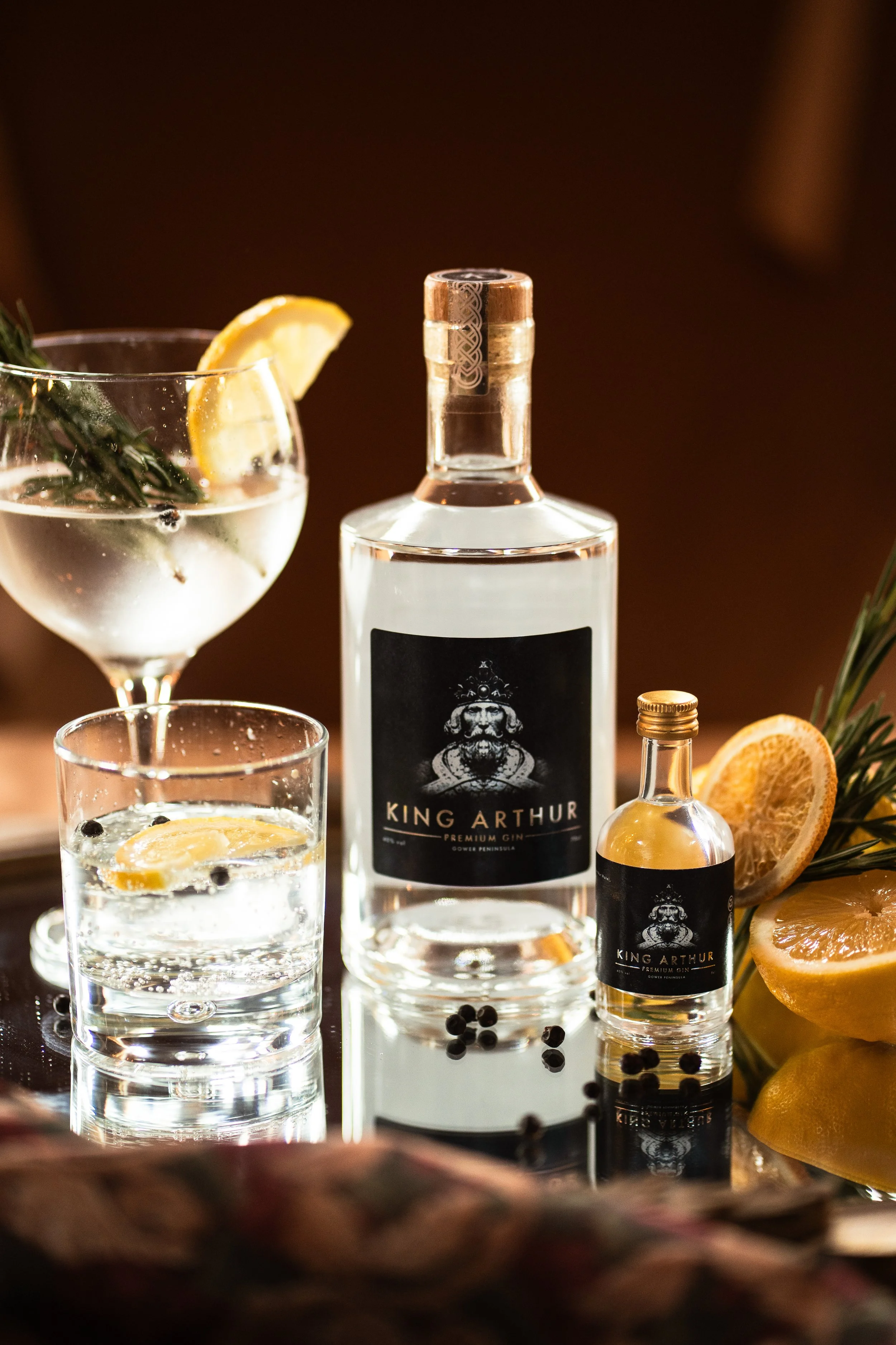A bottle of King Arthur premium gin, a smaller bottle of King Arthur gin, a cocktail glass with a lemon slice and rosemary sprig, another glass with lemon slice, lemon slices, black peppercorns, and a sprig of rosemary.