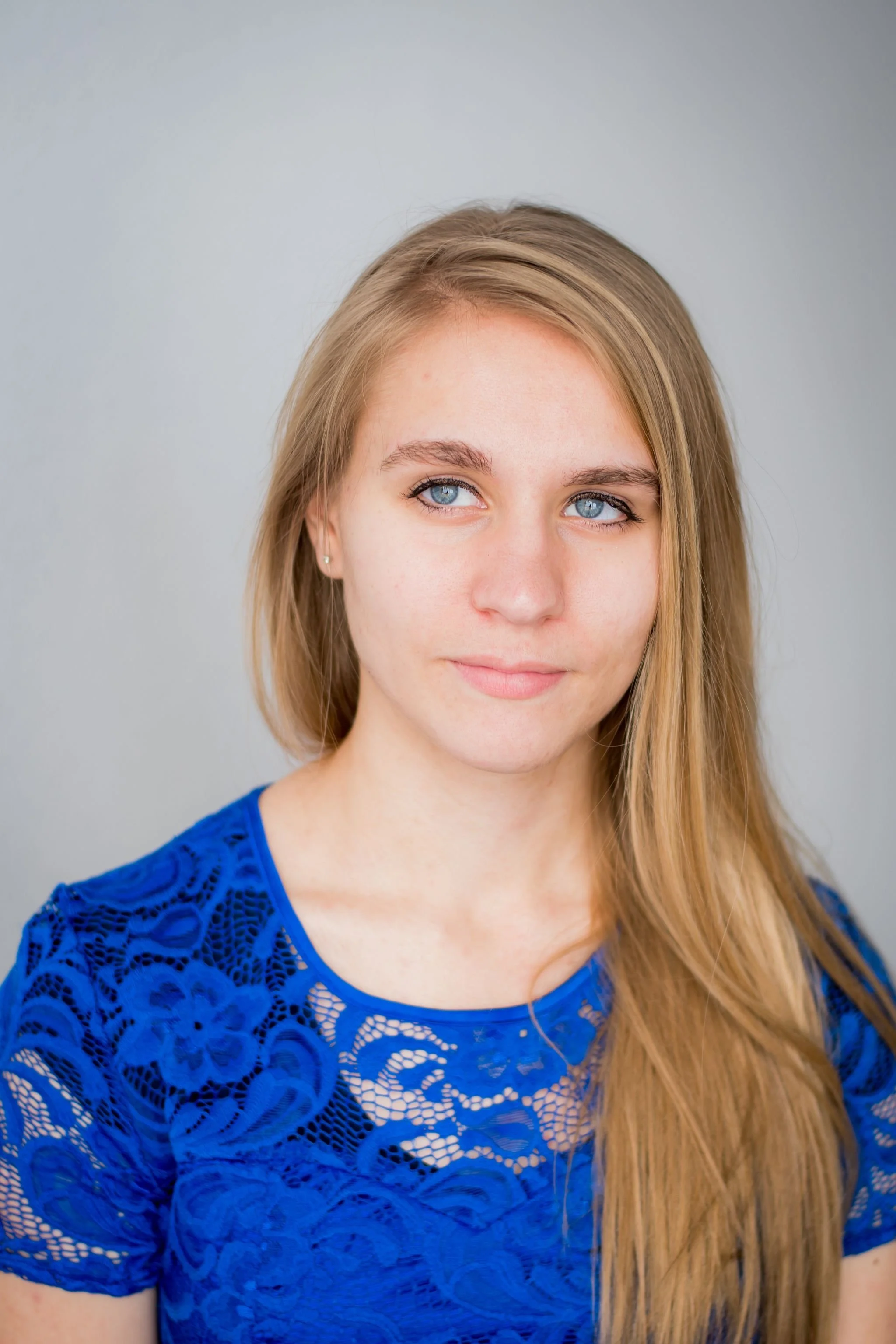 Portrait of a young woman with long, blonde hair wearing a blue lace top, looking slightly to the right with a neutral expression against a light gray background.