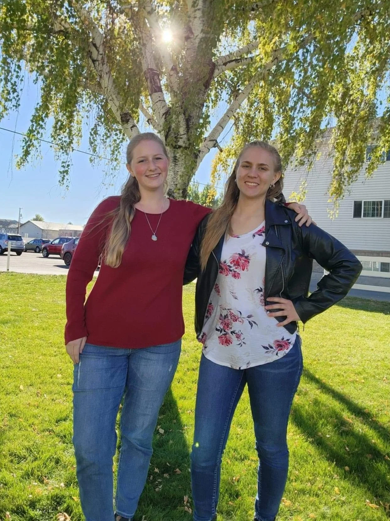 Two women (Emily, left and Sierra, right) standing outdoors in a grassy area under a tree, smiling at the camera. Emily is wearing a red long-sleeve top and jeans, Sierra a floral top with a black leather jacket, also in jeans.