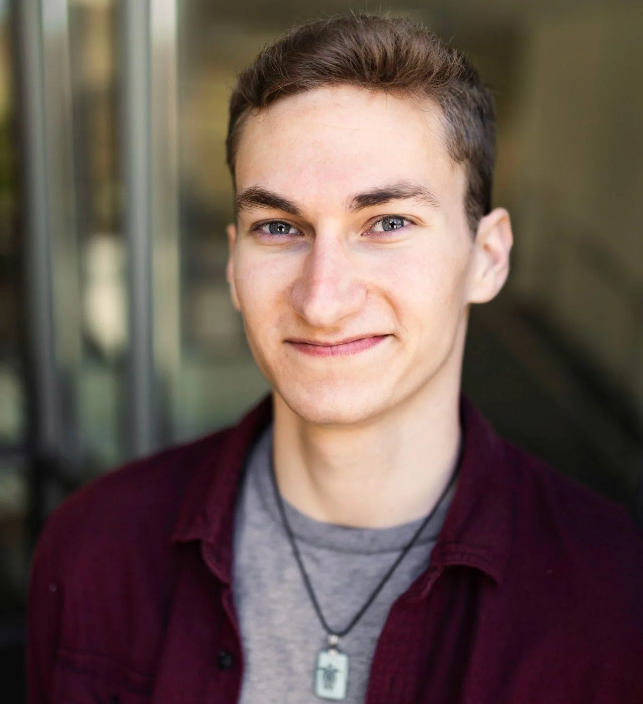 Young man with short brown hair and blue eyes smiling outdoors near a glass building.