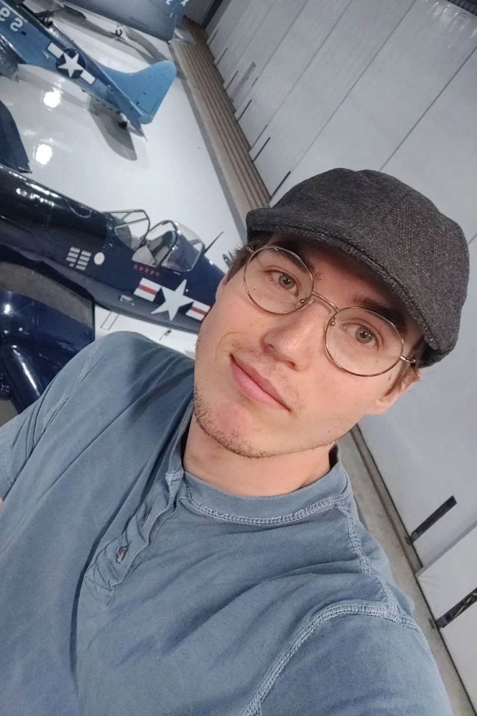 A young man with glasses and a gray flat cap taking a selfie in front of a display of model aircrafts inside a building.