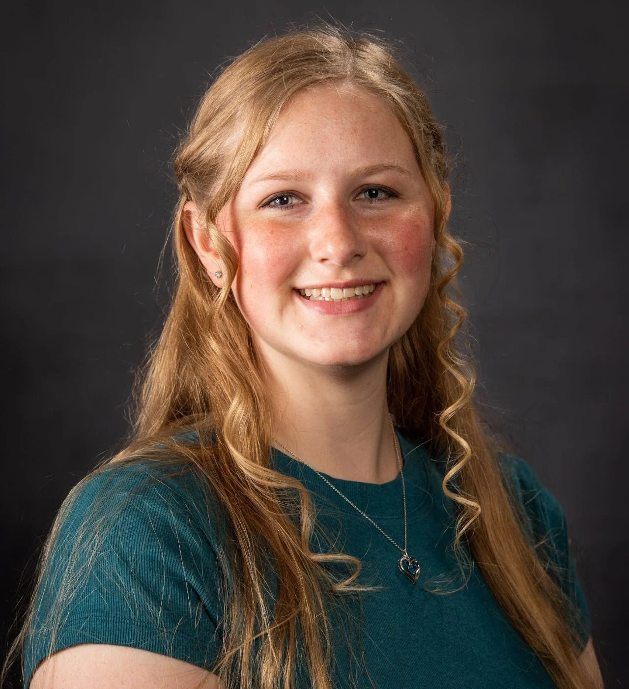A young woman with long, wavy, blonde hair wearing a teal shirt and heart-shaped necklace, smiling in a professional portrait against a dark background.