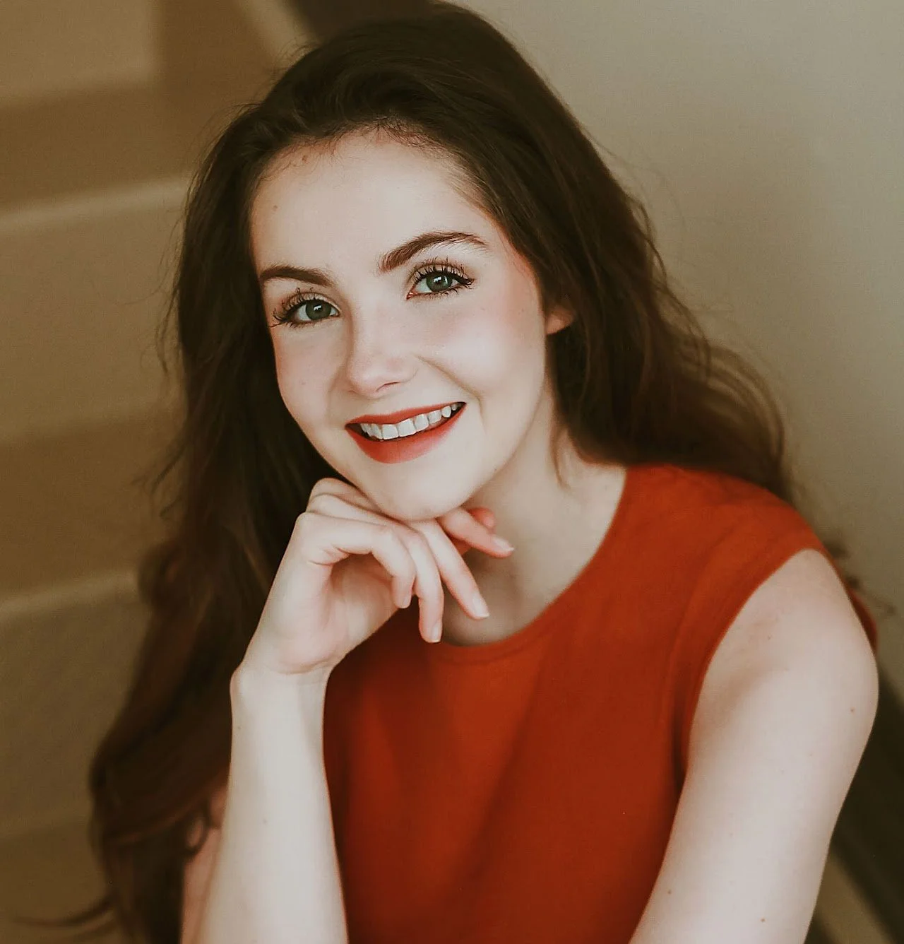 A young woman with long brown hair, green eyes, and fair skin is smiling and resting her chin on her hand. She is wearing a sleeveless red top and has makeup applied, including red lipstick.