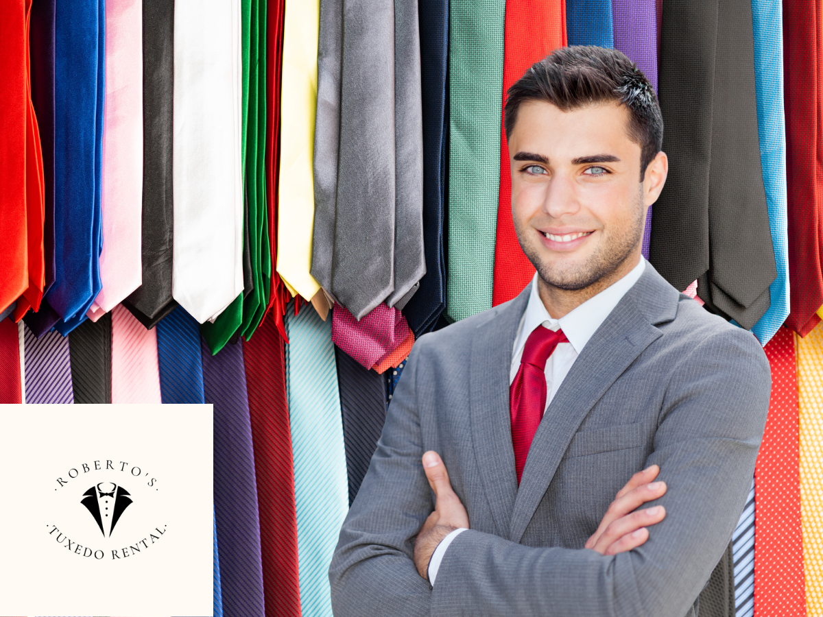 A smiling man in a gray suit and red tie standing in front of a display of colorful neckties at Roberto's Tuxedo Rental.