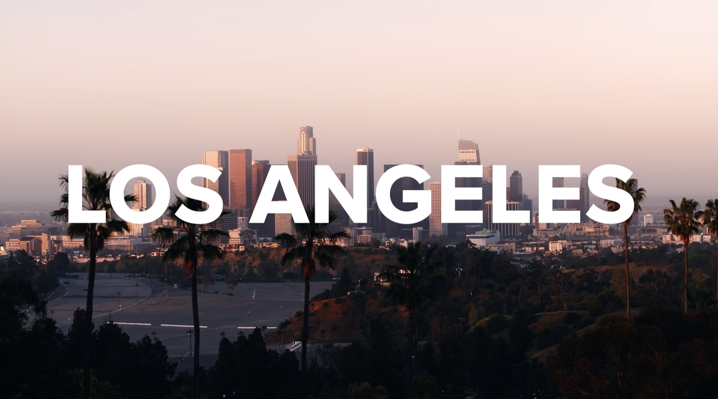 Skyline of downtown Los Angeles with tall buildings and palm trees, overlayed with the text 'LOS ANGELES' in large white letters.