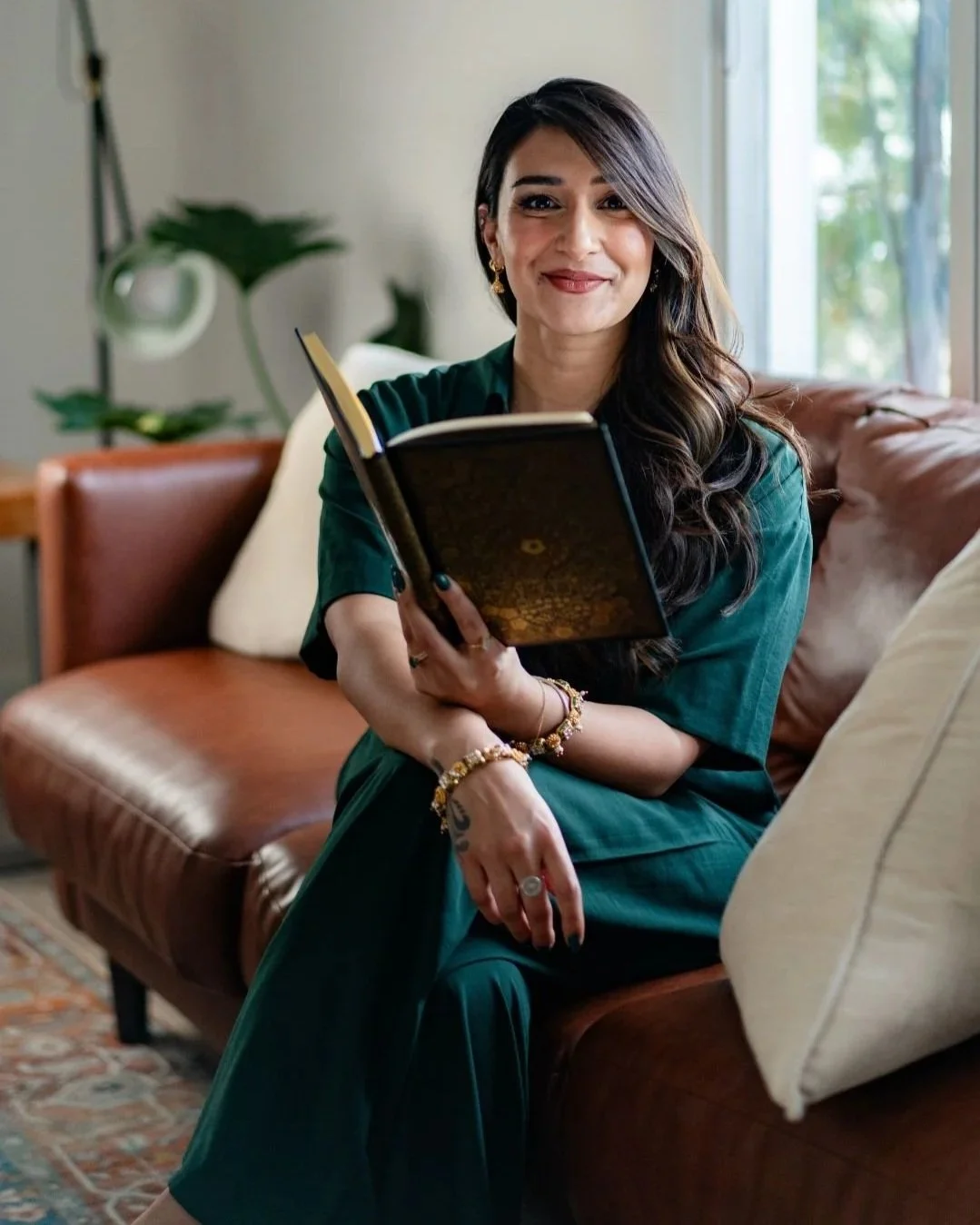 A woman with long dark hair and earrings sitting on a brown leather couch, holding an open book, smiling, with a window and green plants in the background.