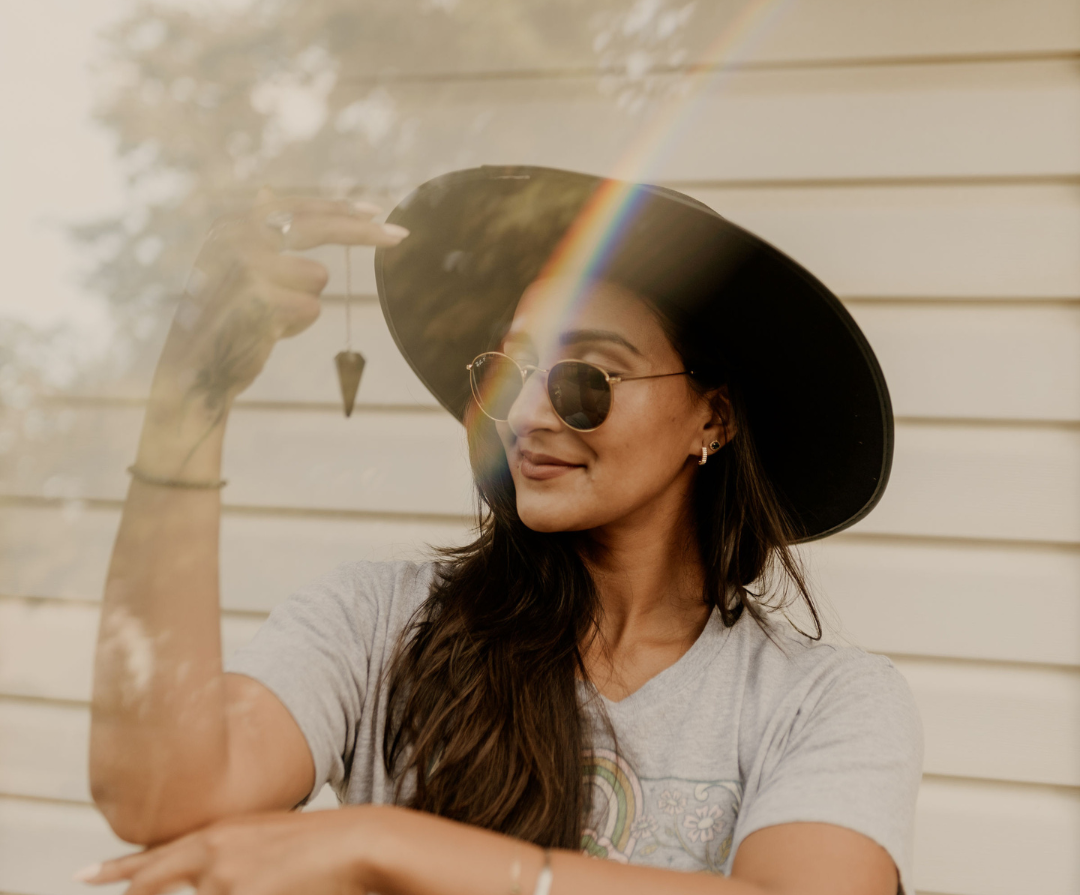 Woman with long hair wearing sunglasses and a wide-brimmed hat, smiling and pointing at a rainbow formed by sunlight reflecting off a mirror.