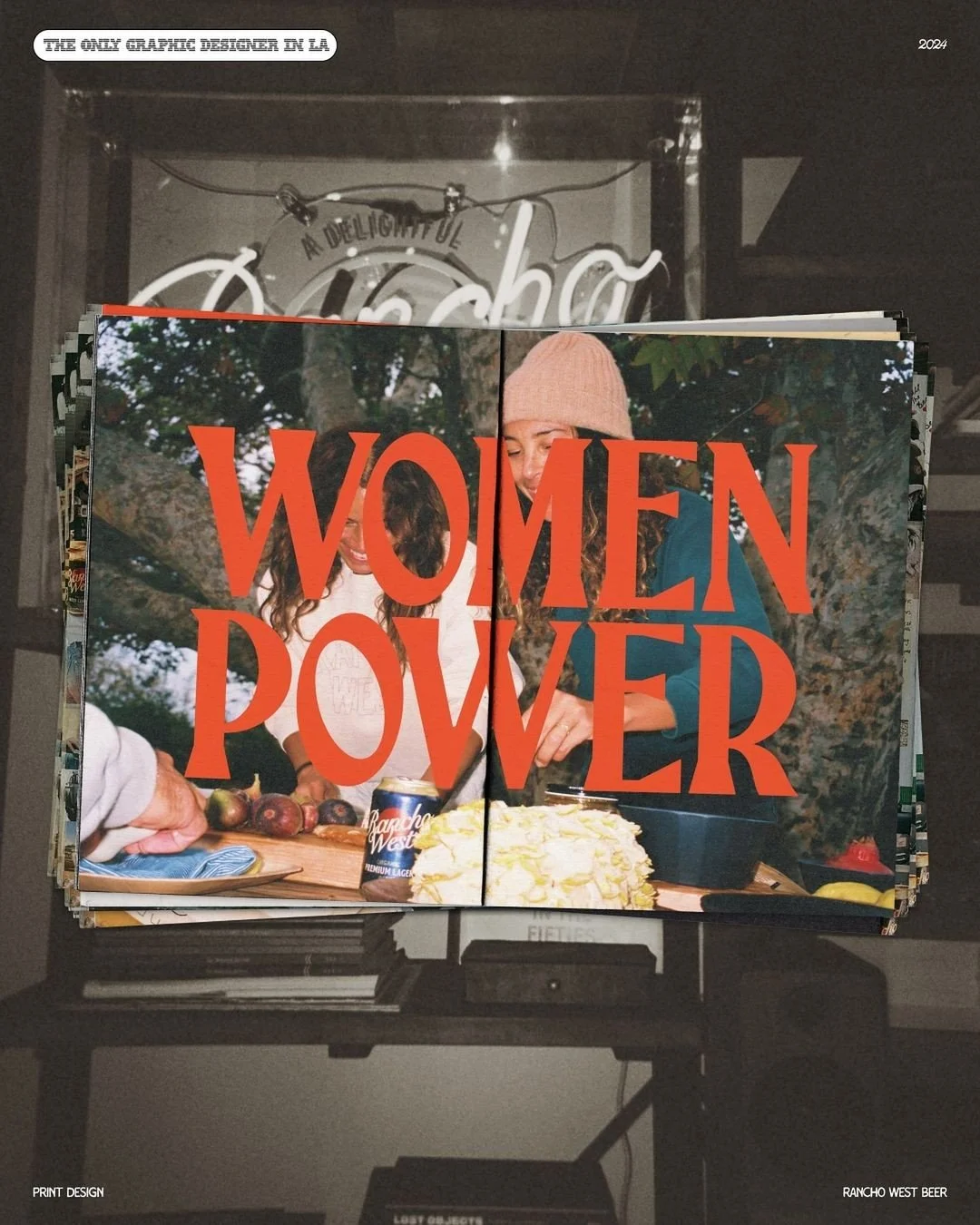 Photo of a magazine or poster with the words 'Women Power' in large red text over a picture of two women outdoors, one wearing a pink beanie. Behind the image, there is a background sign that reads, 'The Only Graphic Designer in LA,' with a neon sign in the shape of a circle above it. The scene appears to be in a cozy, indoor setting, possibly a cafe or store.