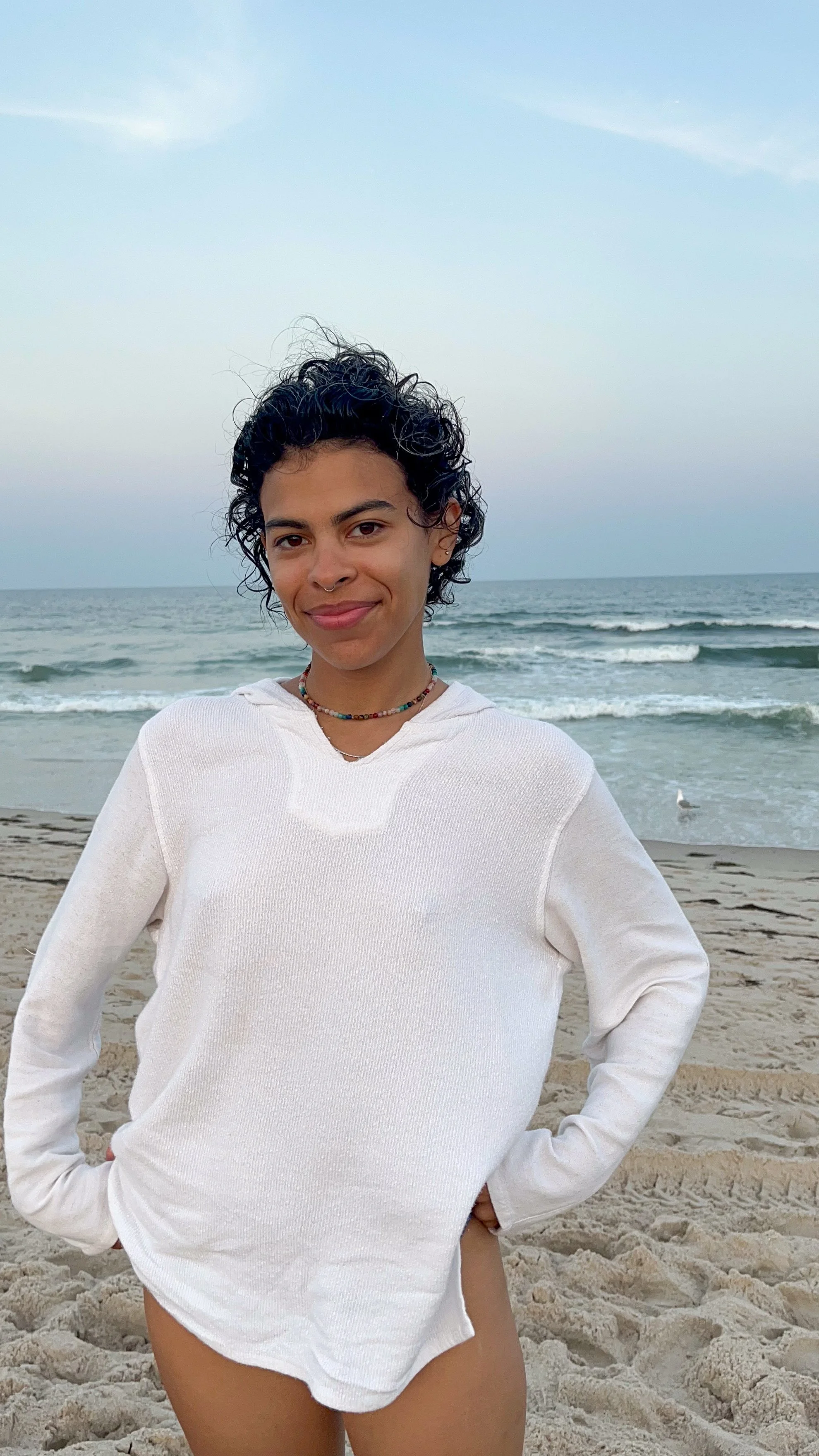 A young woman with curly black hair posing on a sandy beach, wearing a white long-sleeve top and a beaded necklace, with the ocean and sky in the background.