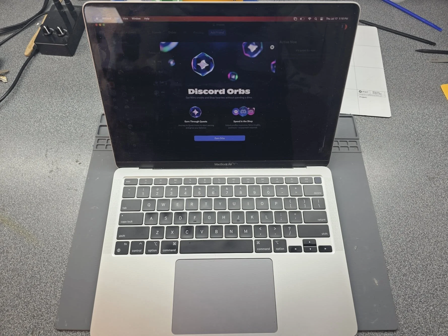 A laptop displaying the Discord Orbs webpage on its screen, placed on a gray desk with various office supplies around it, including a black power strip on the top left, a white notepad, and some black and orange pens.