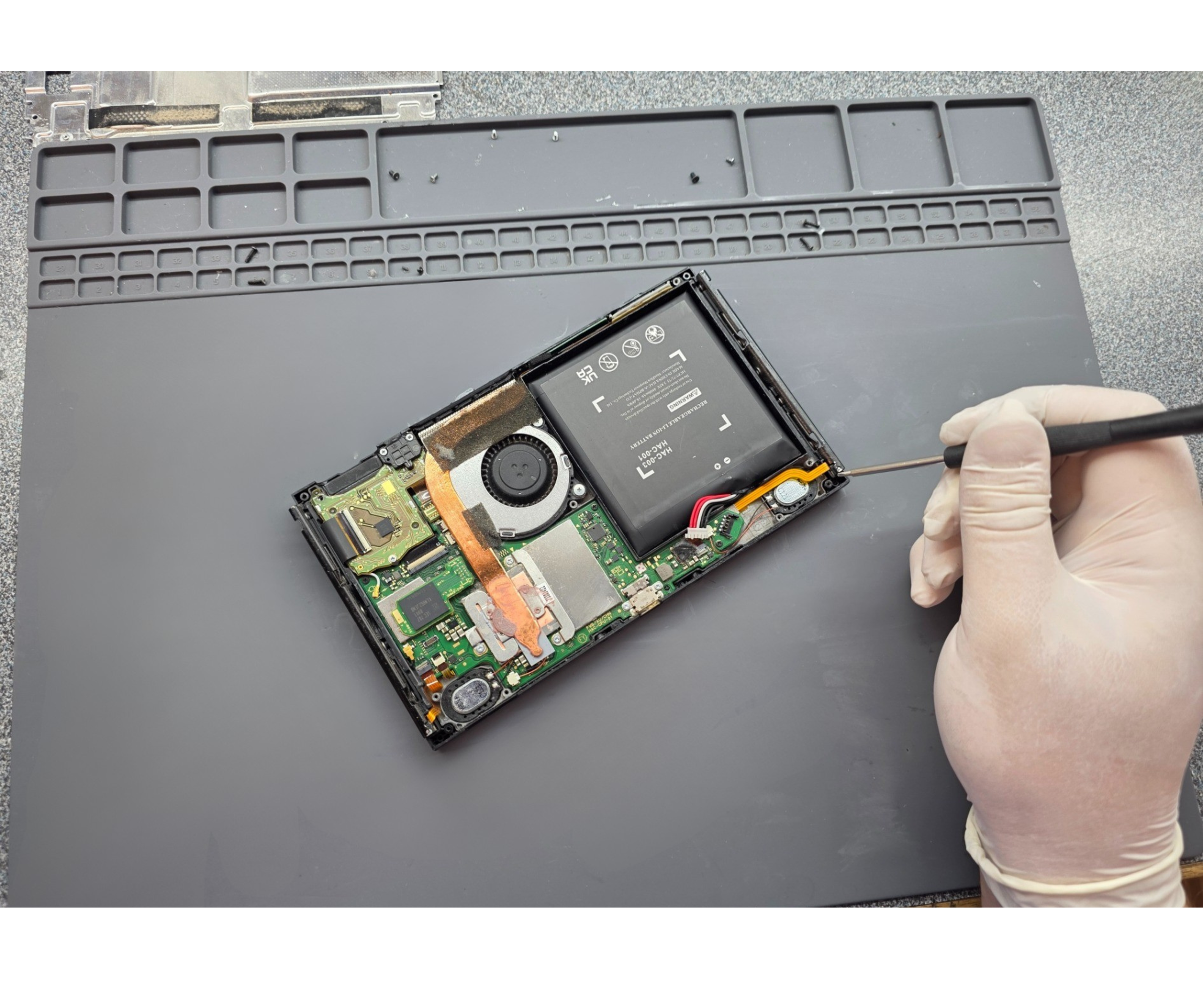 A person wearing latex gloves repairing a tablet by using a screwdriver on the device's internal components, with the back cover removed revealing the battery and circuitry.