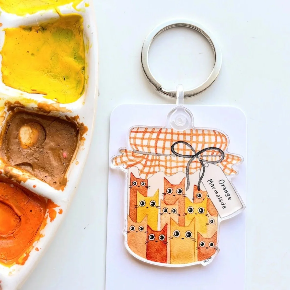 A watercolor keychain art piece featuring a blue owl sitting on water splashes, attached to a white tag with the text "Owlfeathers Watercolor". A watercolor palette with multiple colors is in the upper left corner.