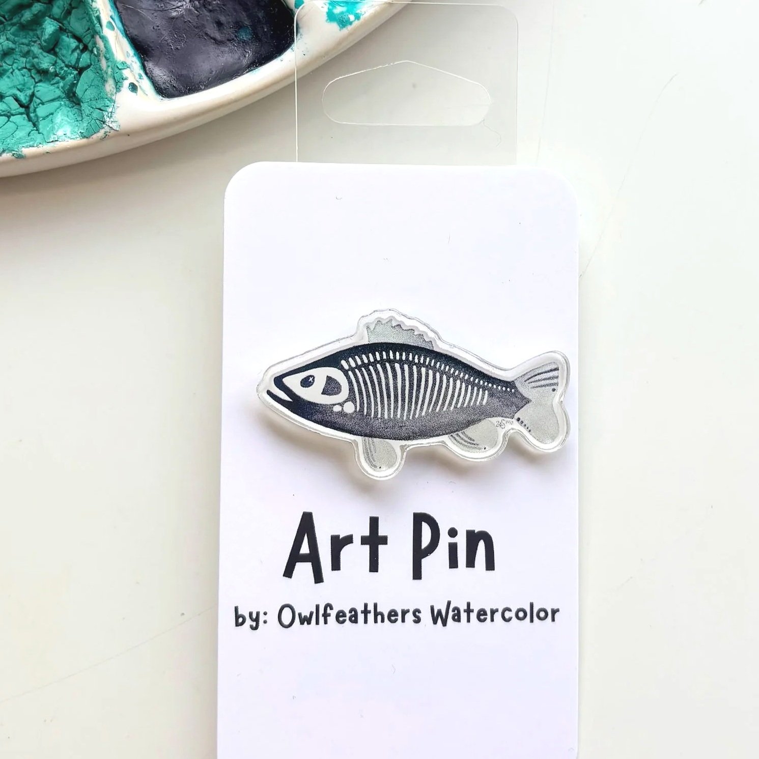 A watercolor paint palette with various colors and an art pin depicting blue cats in a basket, labeled 'Art Pin by: Owlfeathers Watercolor' on a white background.