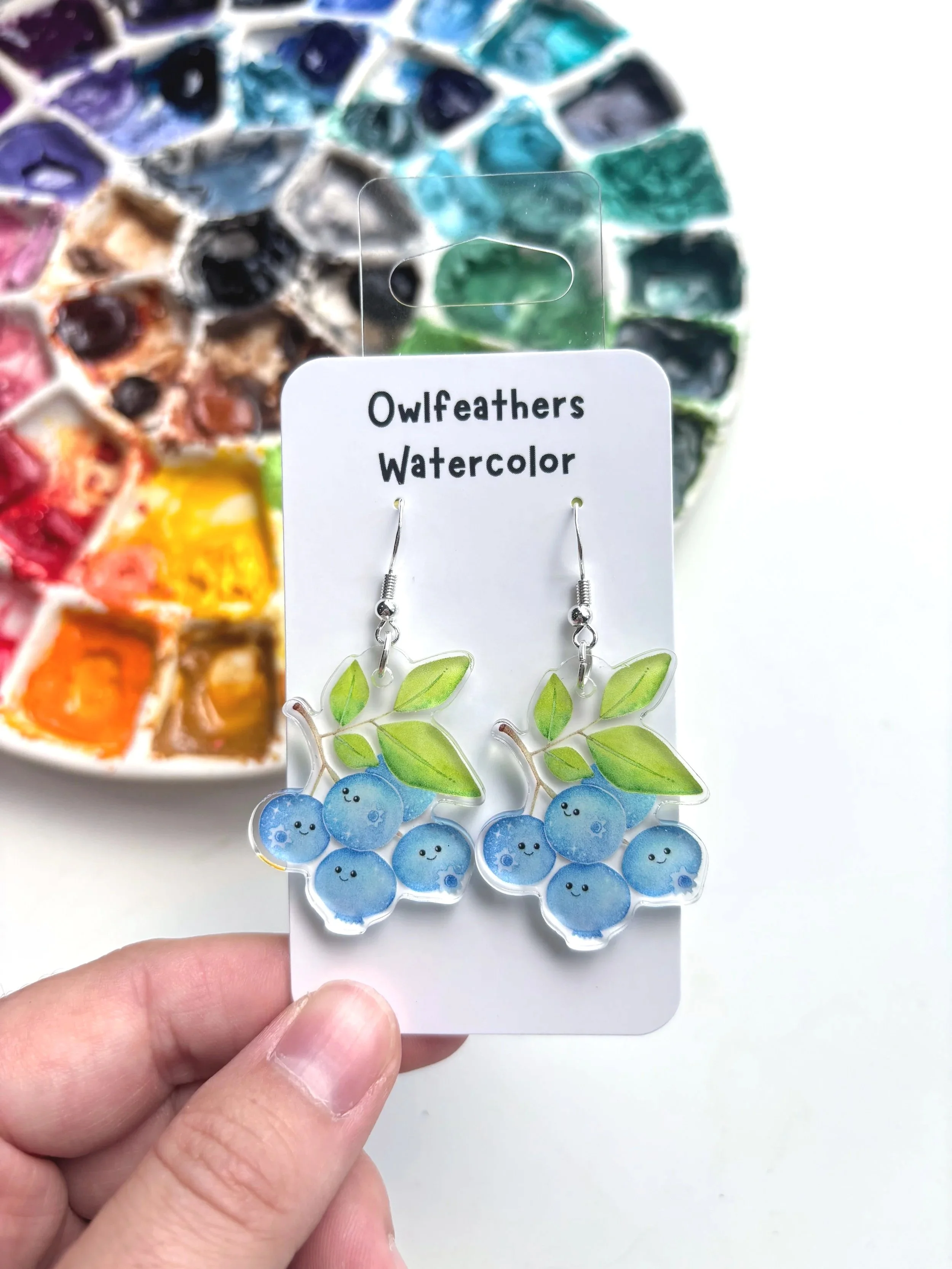 Watercolor earrings shaped like a horse with a soccer ball on a white background, next to a watercolor palette with various shades of red, pink, orange, yellow, and brown.