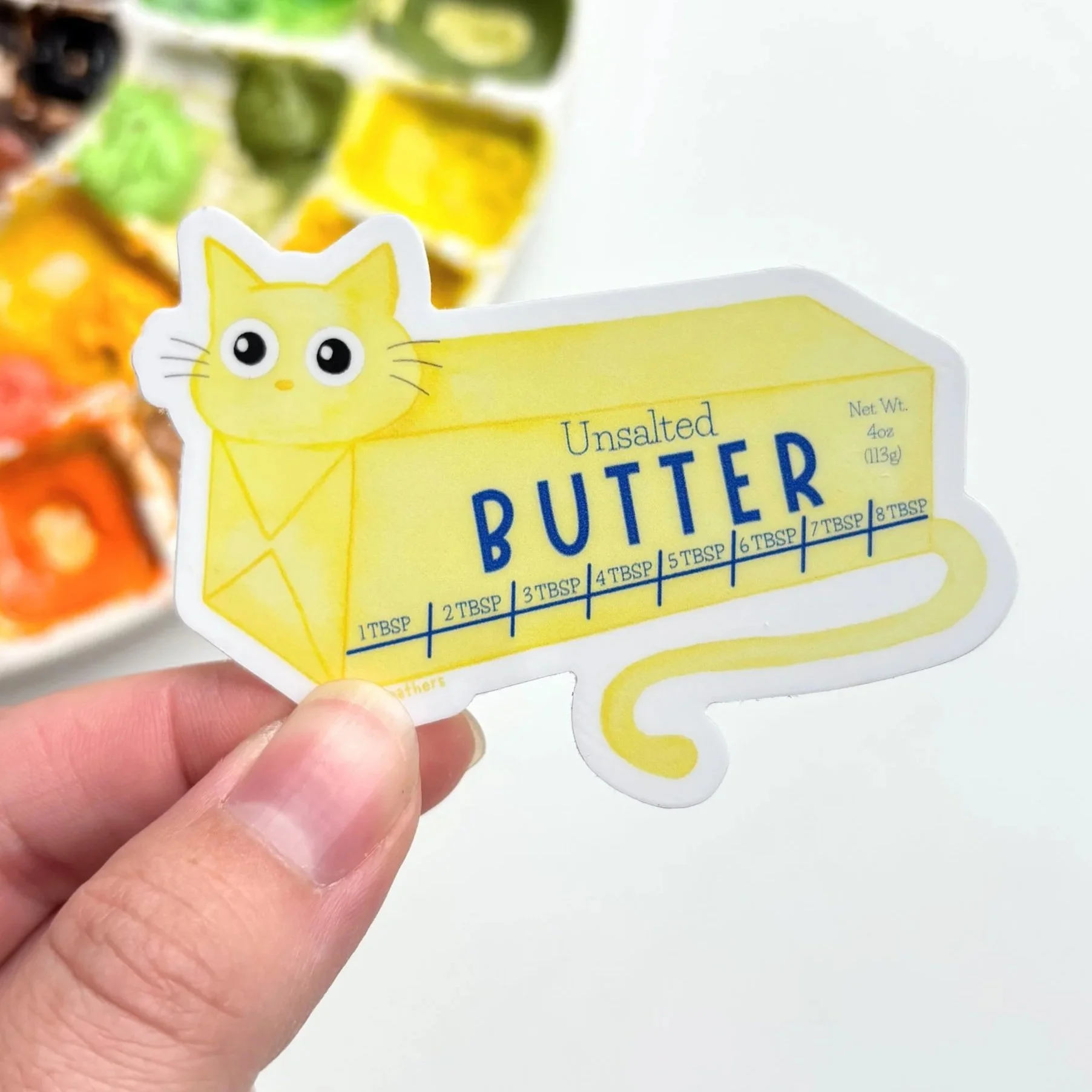 A sticker of a bird with various toy-like illustrations, including a cat, a container of Play-Doh, and some miniature furniture, placed on a white surface next to a paint palette with green, yellow, and blue paints.