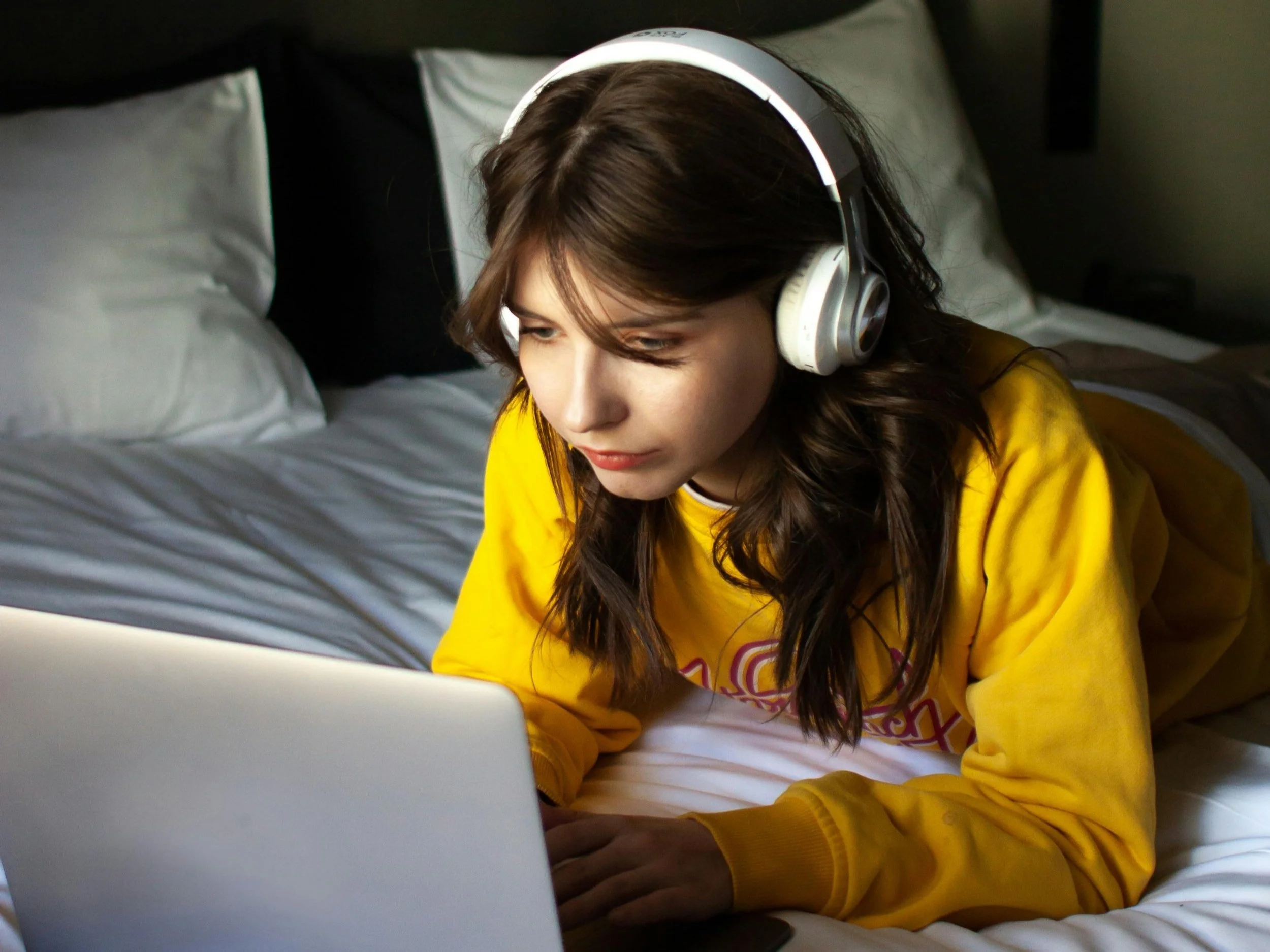 Teen girl lying on a bed with headphones using a laptop for online therapy.