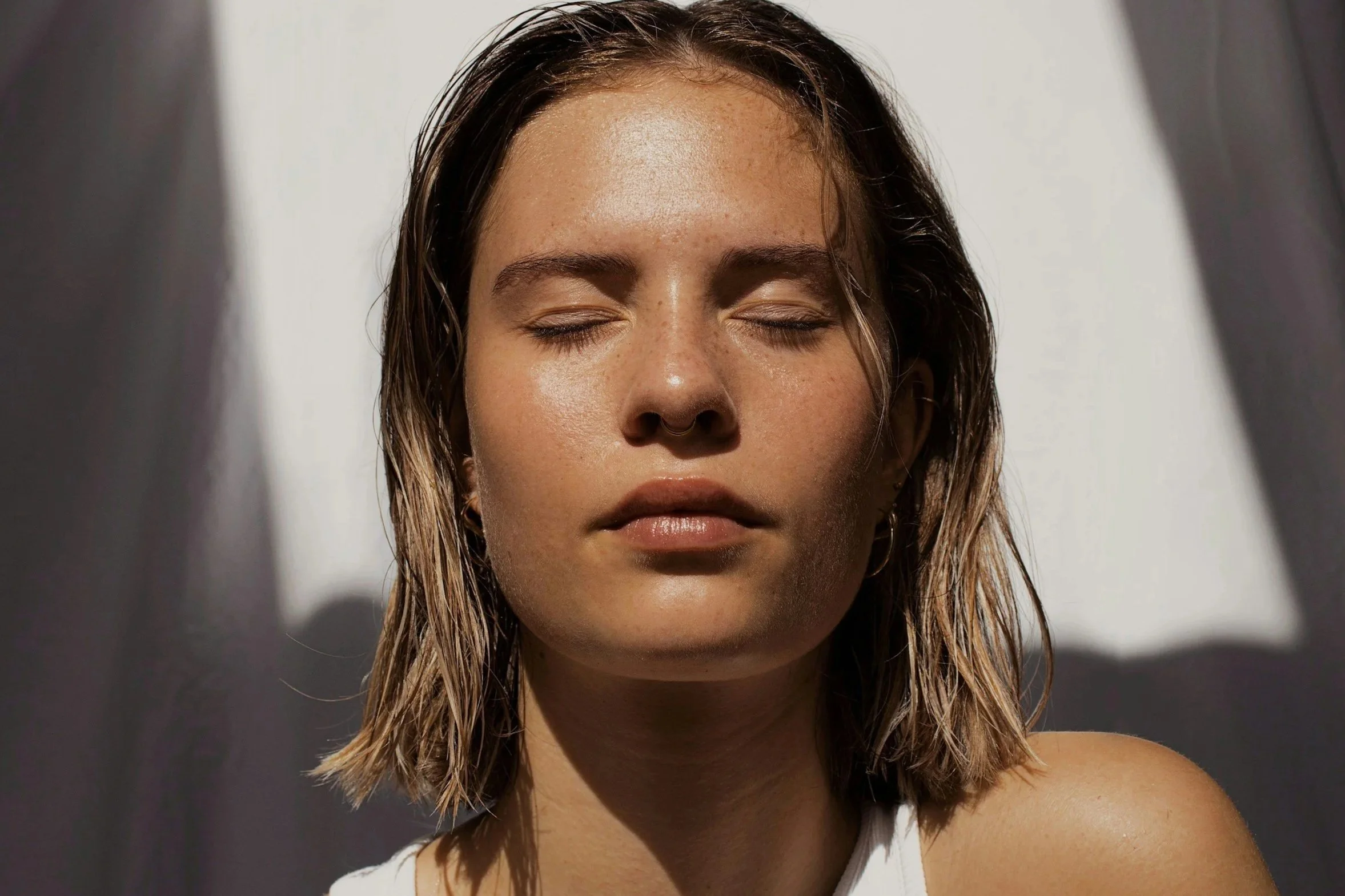 Close-up of a woman with closed eyes and wet hair, sitting with sunlight casting shadows on her face.