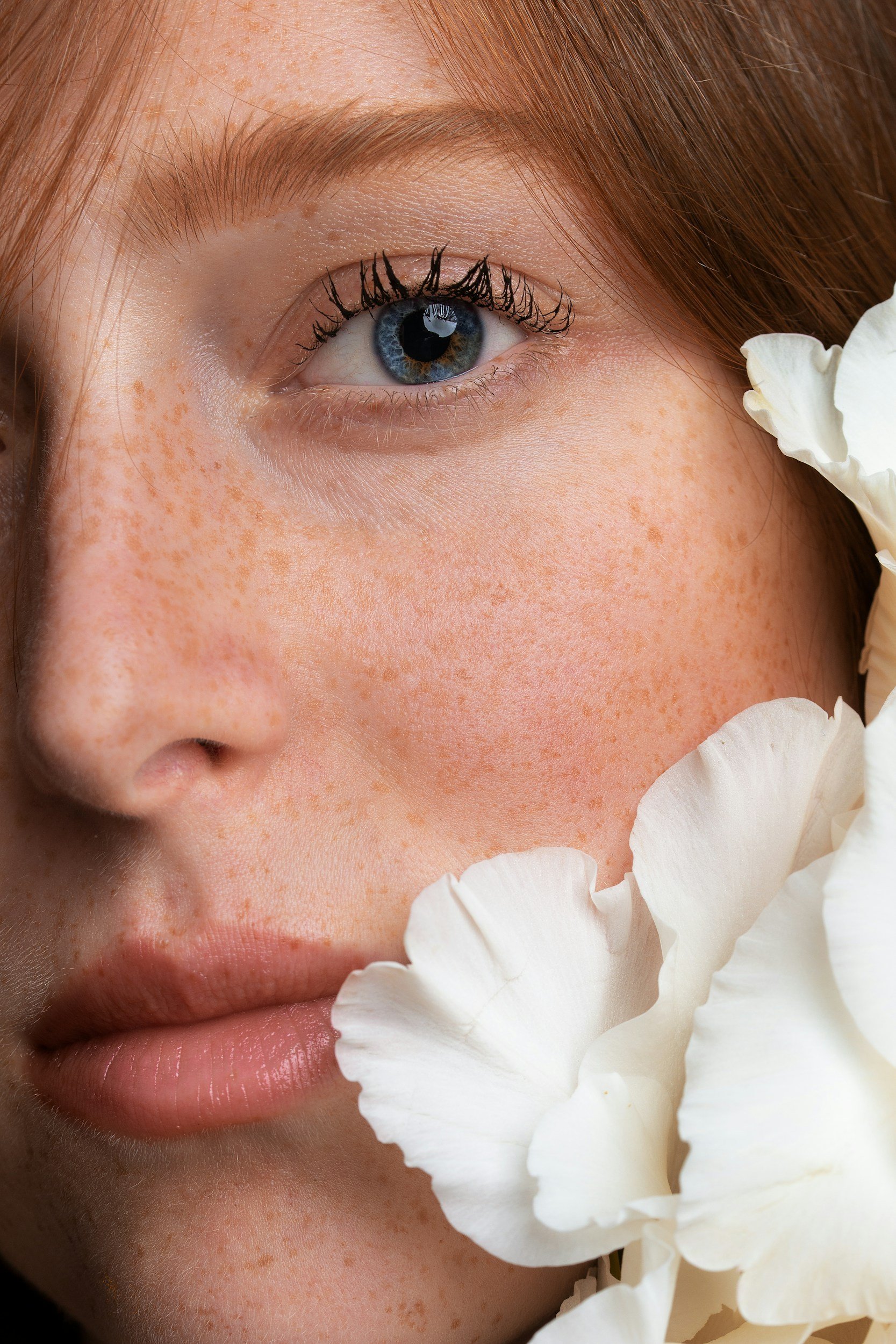 Close-up of a young woman's face with freckles, showing her left eye, nose, and lips, with white flowers near her cheek.