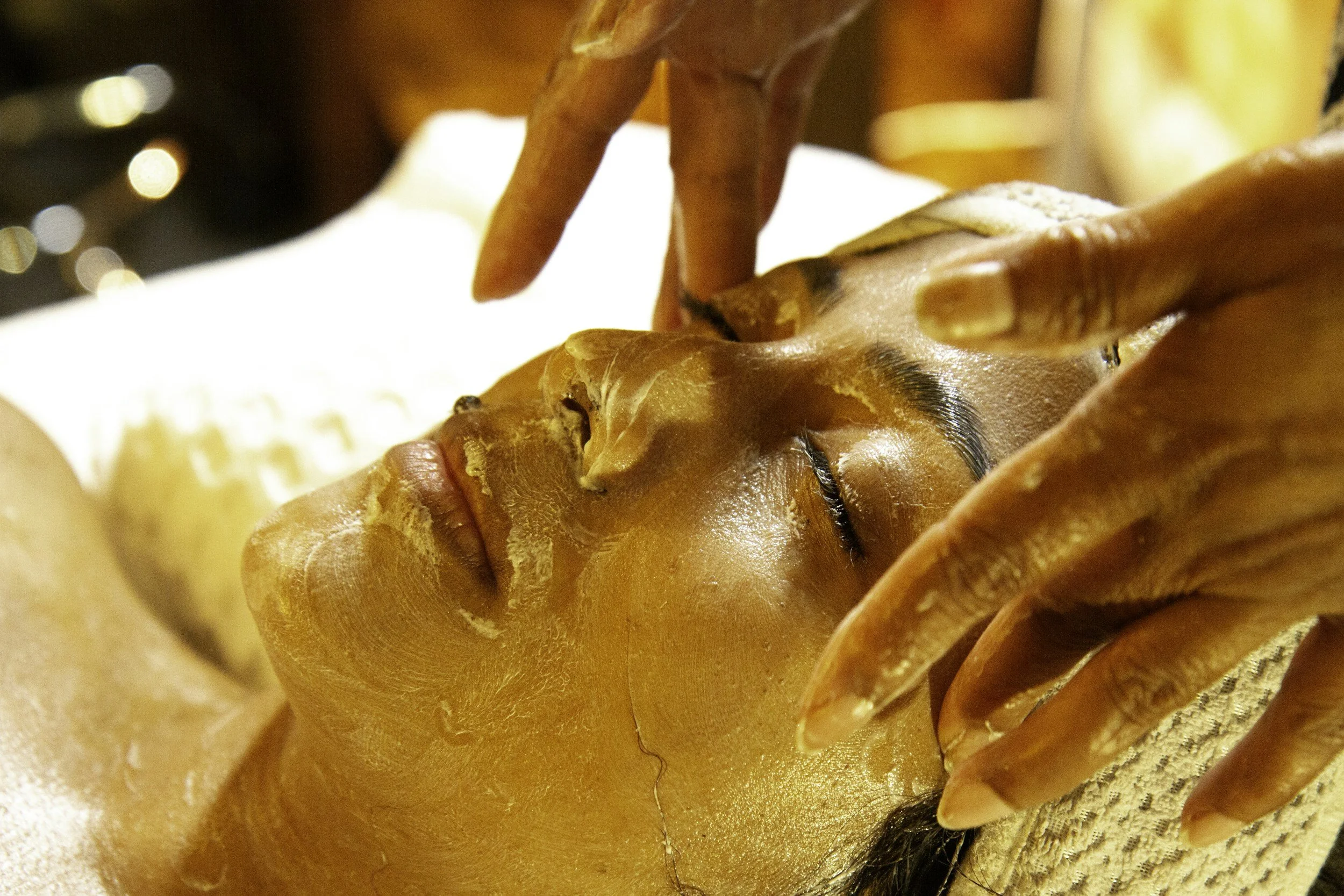 A person receiving a facial massage in a spa setting, with their eyes closed, lying on a treatment bed covered with a towel.