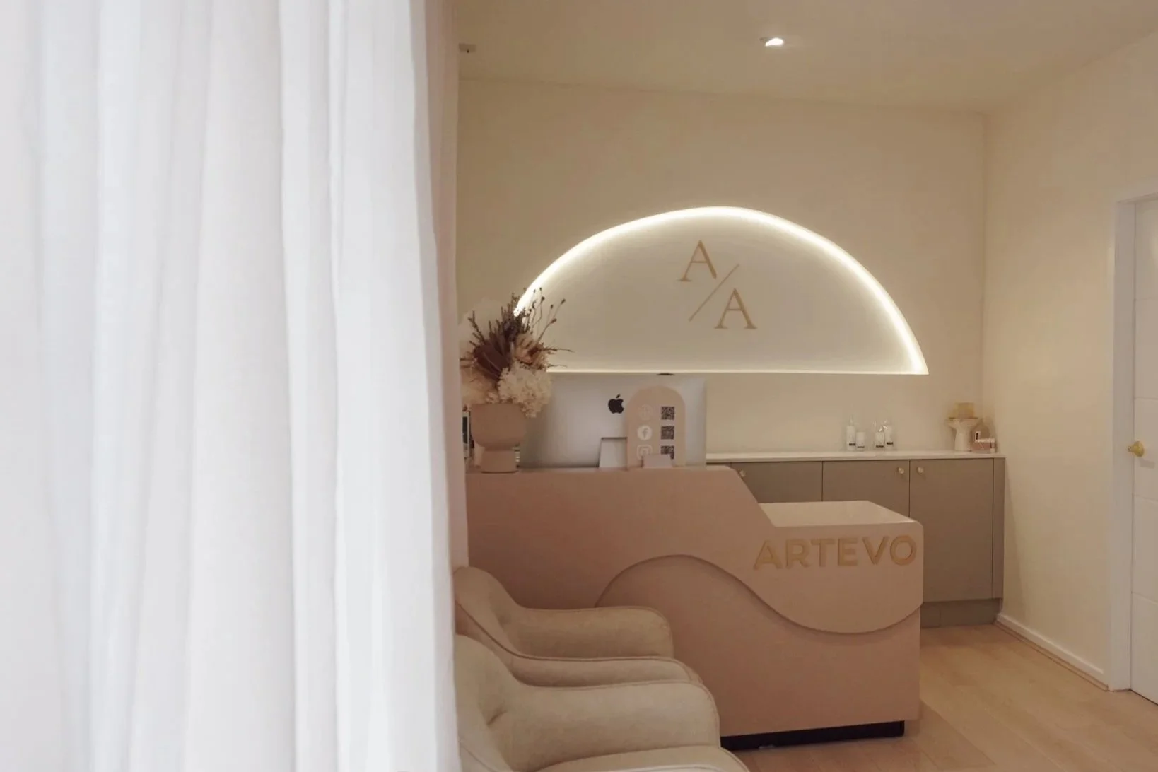 A reception area with a beige curved desk that has the word 'ARTEVO' in gold letters, a computer monitor, a vase with dried flowers, and a beige armchair. A semicircular wall feature with backlit lighting and gold 'A/A' initials is visible behind the desk, along with a gray cabinet and several small bottles or jars on top.