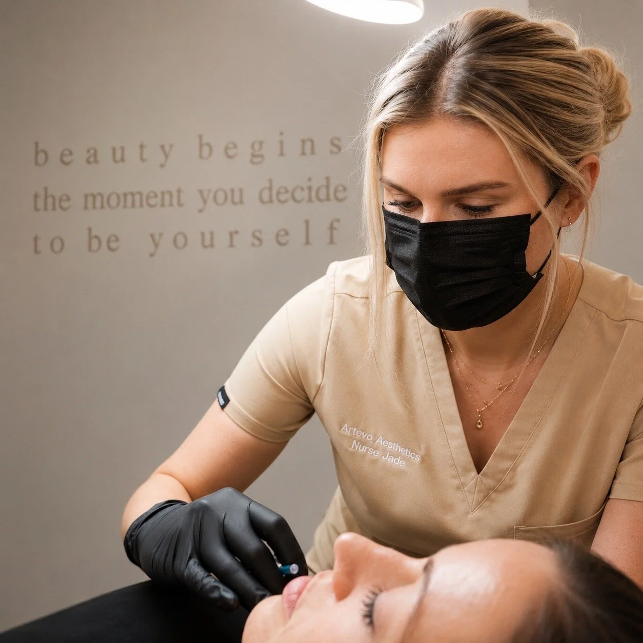 A woman with blonde hair wearing a black mask and black gloves is performing a medical or cosmetic procedure on a woman lying down. The woman receiving the treatment has dark hair and is resting with eyes closed. In the background, there is a wall with a motivational quote that says, 'Beauty begins the moment you decide to be yourself.'