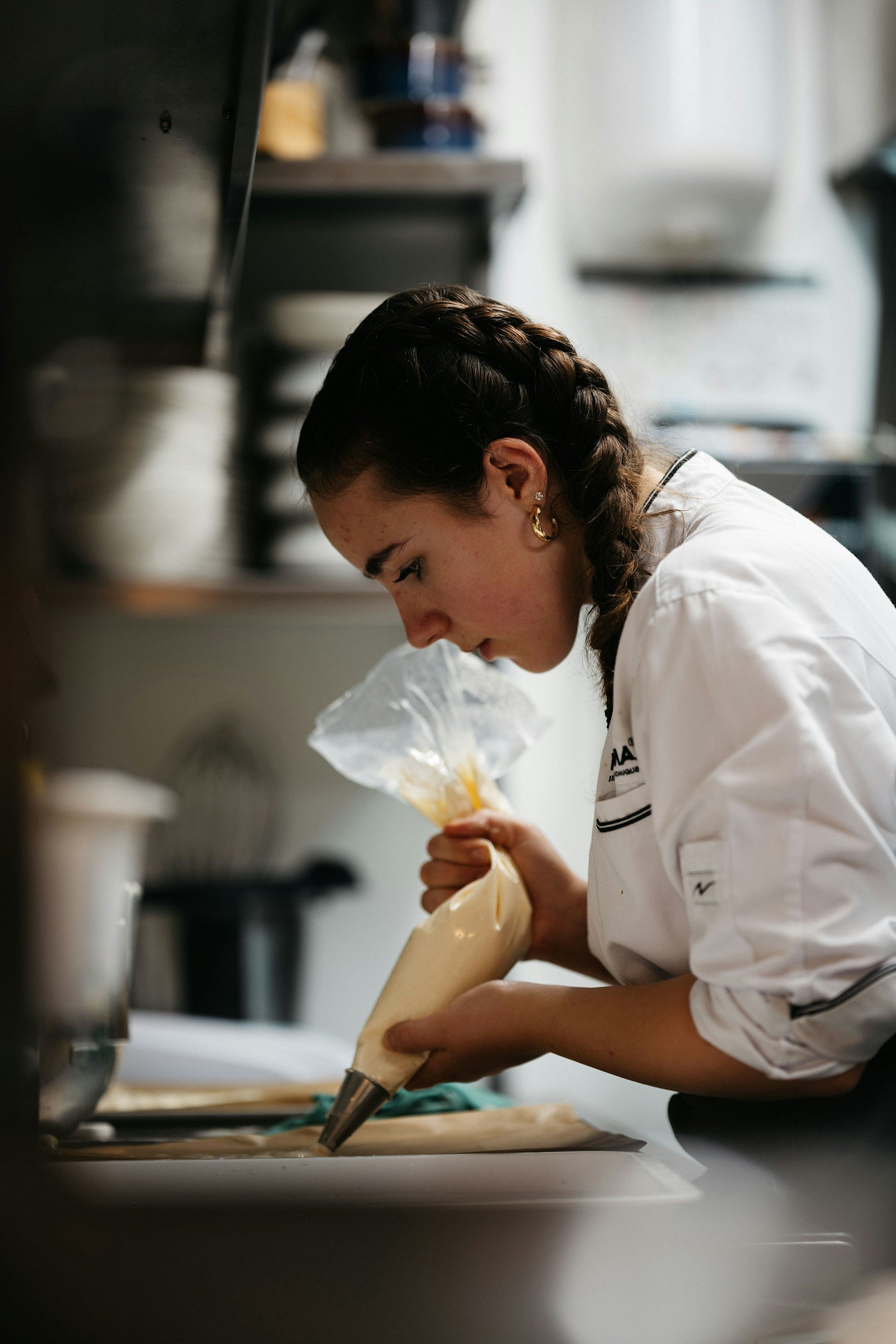 A female chef with dark hair in braids, wearing gold hoop earrings and a white chef's coat, focuses on piping cream or batter from a plastic bag onto a surface in a professional kitchen.