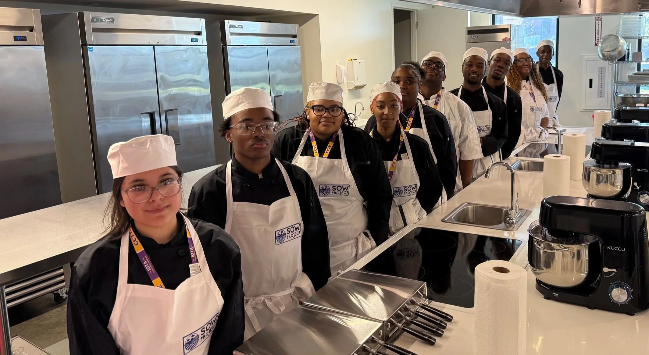 Group of young people dressed as chefs in a kitchen classroom, standing in line behind a counter with cooking utensils and appliances, wearing aprons and chef hats.