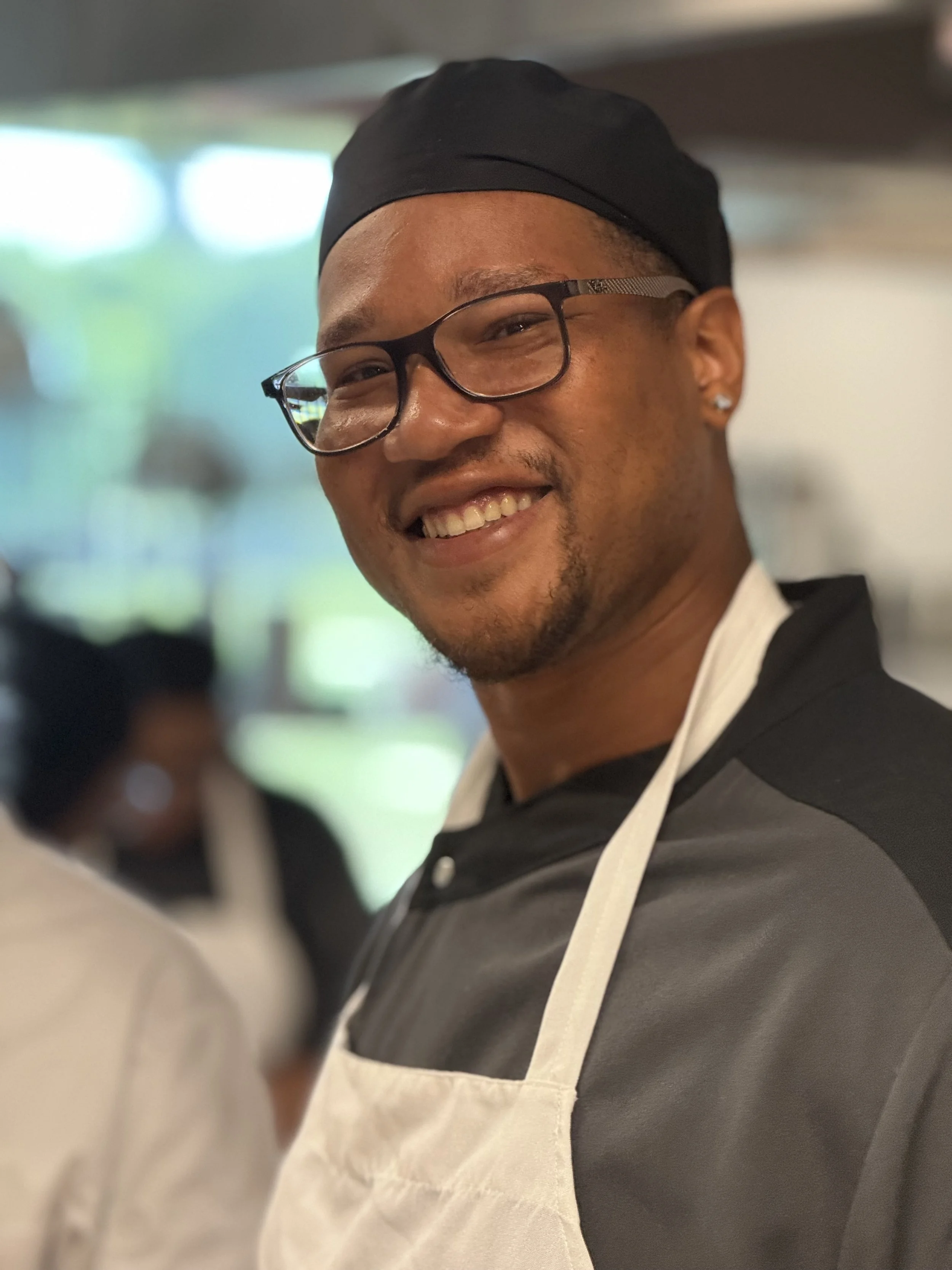 Close-up of a smiling man wearing glasses and a black hair cap, dressed in a black and white apron, possibly working in a kitchen or restaurant.