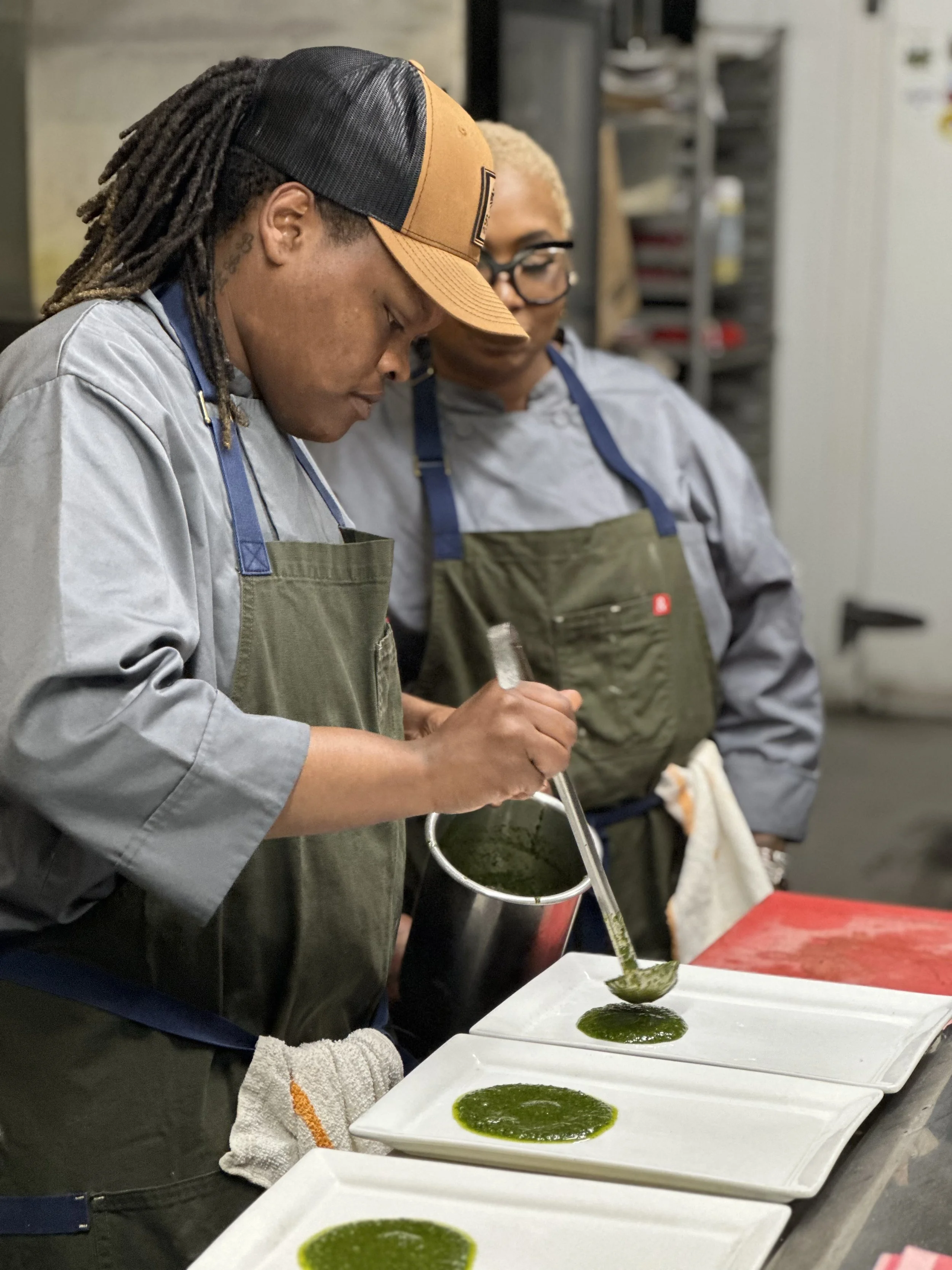 Two chefs, wearing gray jackets and aprons, preparing green sauce in a kitchen. One chef is pouring the sauce onto plates, while the other watches. Three plates with evenly spread green sauce are on the counter.